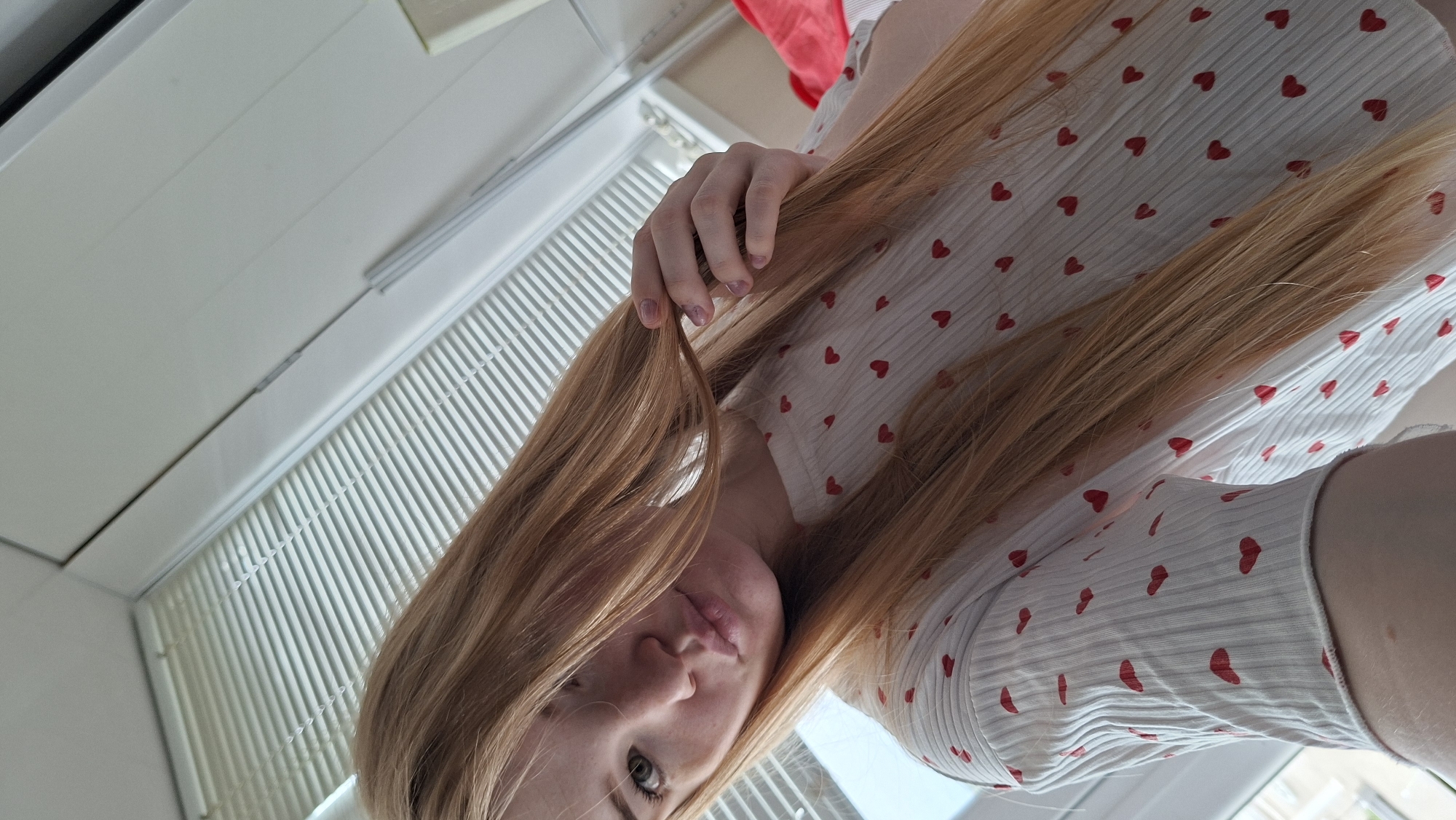 A photograph taken in an indoor location within the city of Chaikovsky, Russia. The photo is taken from a low angle looking upwards at a woman. The woman has fair skin and light brown hair, wearing a white shirt adorned with small red hearts. The photograph includes a section of a window with white blinds. The details provided are based solely on the visual information within the image.