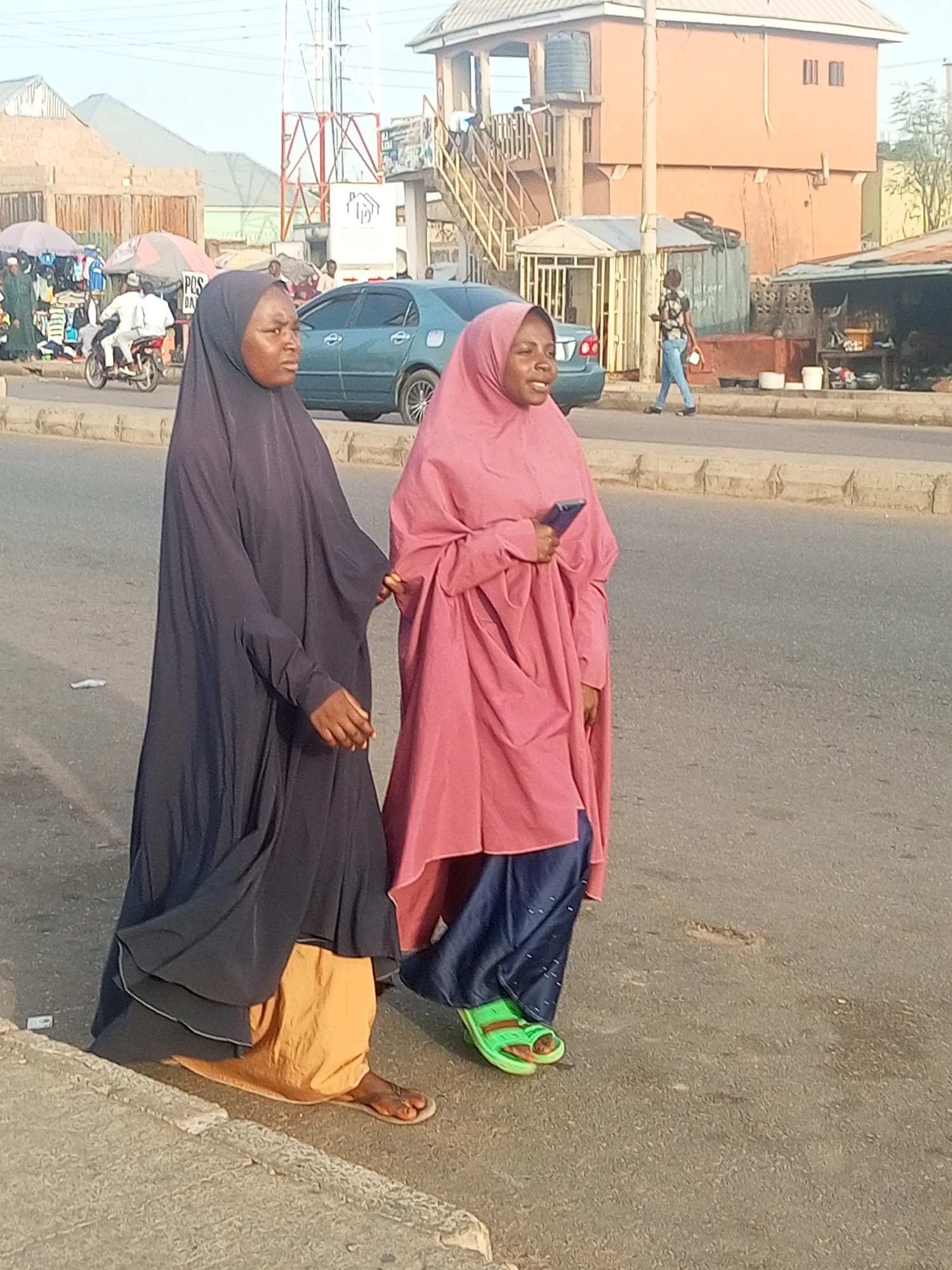 Two women wearing hijabs are walking on a street in Tempure, Nigeria. The woman on the left is wearing a dark blue hijab and a yellow skirt with sandals. The woman on the right is wearing a pink hijab with blue pants and green sandals. They are walking next to each other, with the woman on the left holding the woman on the right's hand. The woman on the right is holding a phone. In the background, there is a car, a building, and a pedestrian overpass. Other people are present on the street.