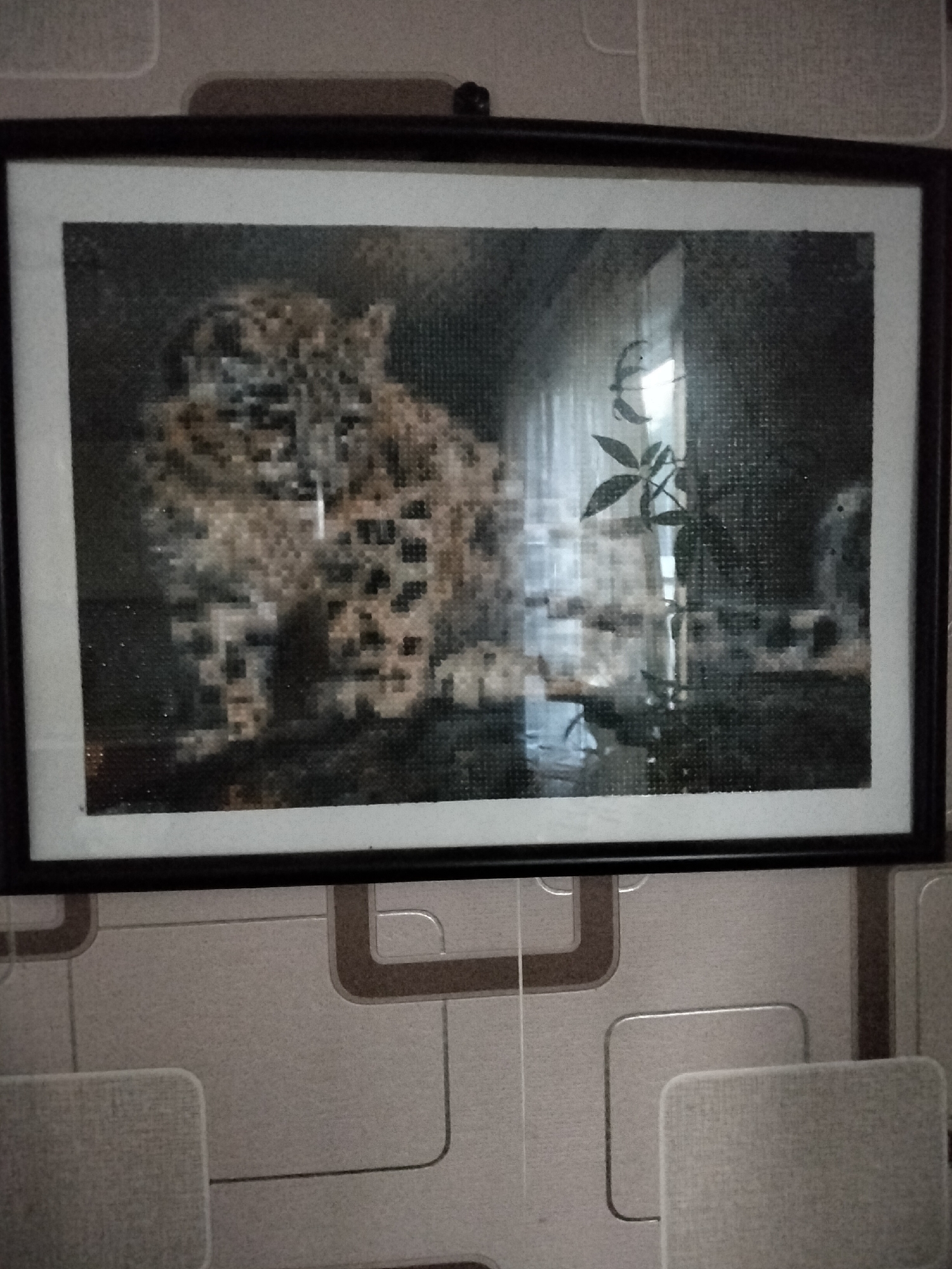 A framed picture of a leopard is hanging on a wall. The leopard is depicted in a diamond painting style, with a focus on its spots and facial features. In the background of the painting, there is a blurry view of a room with a window and curtains, and a plant is visible to the right of the leopard. The frame of the painting is black, with a white matting surrounding the artwork. Below the framed picture, the wall is covered in patterned wallpaper. There are two light-colored chairs visible in the lower part of the image. The image was taken in Nizhnedonskoy, Russia.