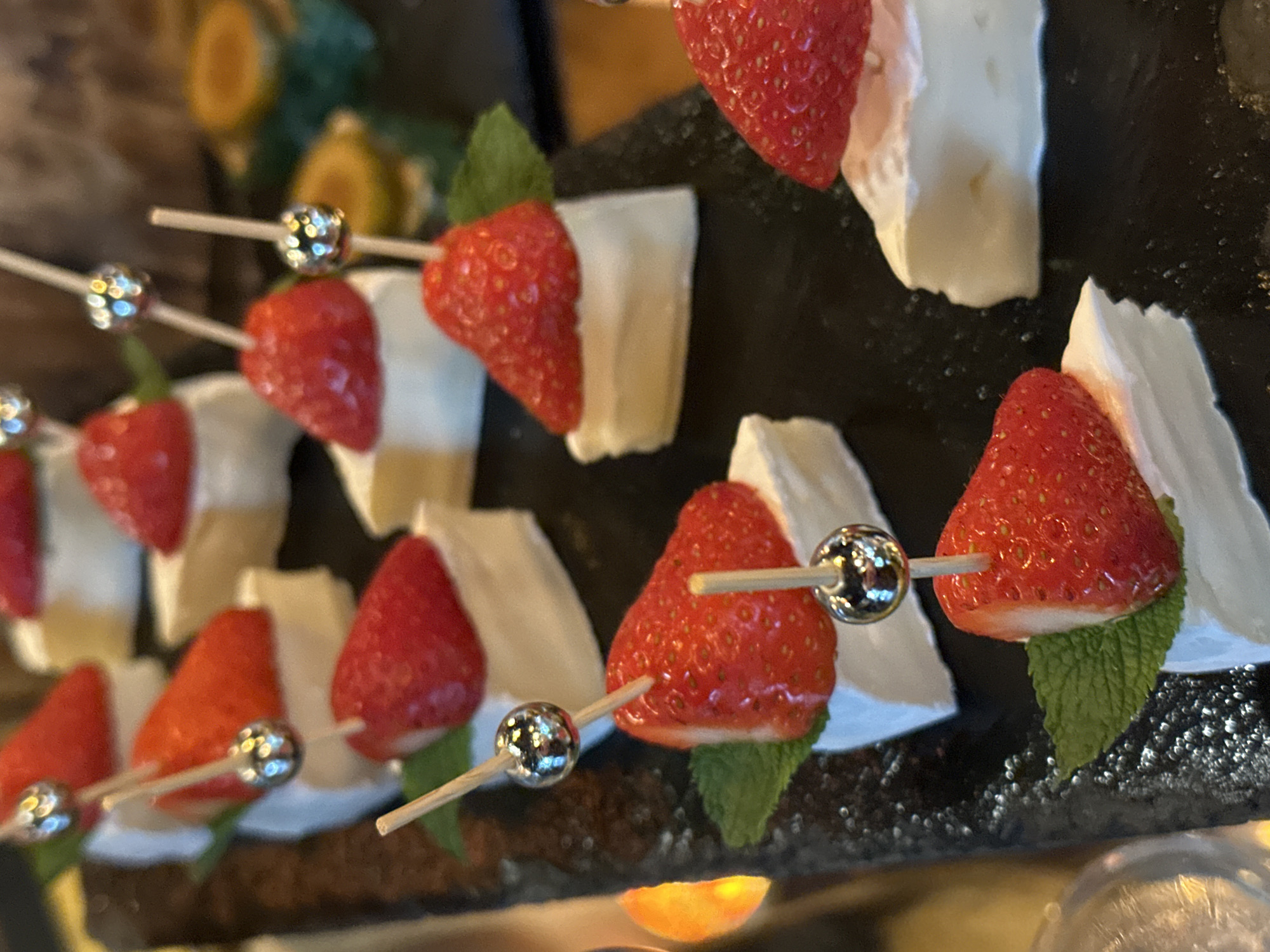 The image shows a platter of appetizers, likely in a restaurant or catering setting. Each appetizer consists of a piece of white cheese (possibly Brie or Camembert), a fresh strawberry, and a small mint leaf. The strawberry and cheese are pierced by a small skewer, some of which are decorated with a small silver ball on the end. The appetizers are arranged on a dark, possibly slate, platter. Based on the information provided, I cannot give you a specific location for this scene in the city of Новоглаголево, Russia.