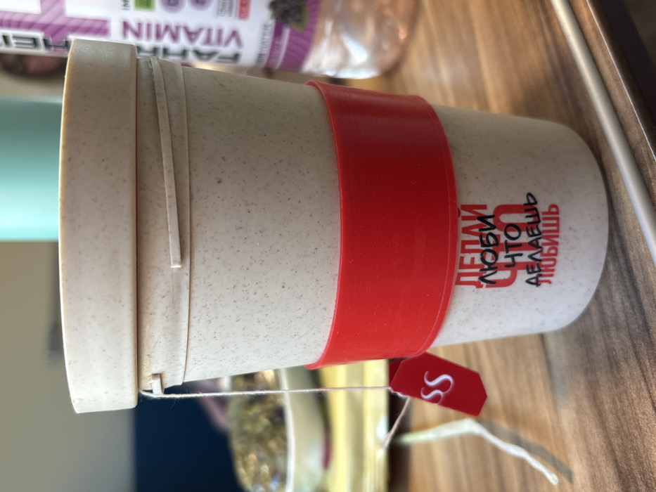 A beige, speckled reusable cup with a matching lid is centrally positioned on a light-brown wooden table. A red silicone band encircles the cup's middle. Below the band, Russian text is printed: "ДЕЛАЙ ЛЮБИ ЧТО ДЕЛАЕШЬ ЛЮБИШЬ," which translates to "DO WHAT YOU LOVE LOVE WHAT YOU DO." A white string extends from a slot in the lid, connected to a small red tag featuring white "SS" lettering, indicating a tea bag. In the blurred background to the left, a white bowl containing dark, textured contents is visible. To the right, a clear plastic bottle with a purple label partially displays "FAIRTRADE VITAMIN WATER" and an image of dark berries. A faint, indistinct reflection of a human figure is discernible behind the bottle. A thin, light-colored rod rests on the table surface in the lower right foreground. This scene is located in Krasnoufimsk, Russia.
