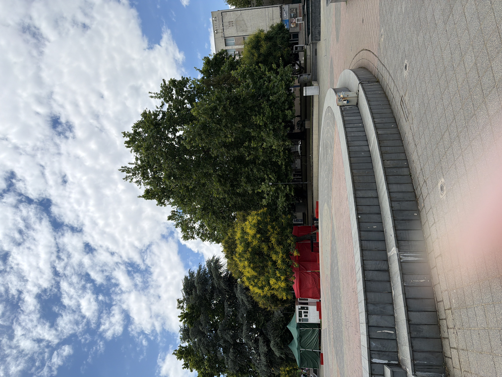 The image captures an outdoor public space in Pazardzhik, Bulgaria. In the foreground, a tiered, circular stage structure made of stone and concrete is visible. The stage is bordered by paving stones. Behind the stage, several large trees provide shade. To the left, two green tents and a small white structure with windows are set up, followed by a larger red tent. In the background, a multi-story building with a light-colored facade stands. The sky above is partially cloudy with a mix of blue sky and white clouds. The scene appears to be a public square or park area, possibly set up for an event or market. No people are clearly visible in the image.