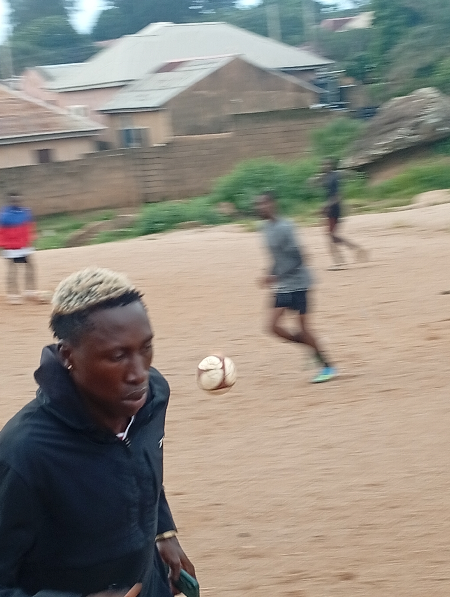 The image depicts an outdoor scene in a sandy area, possibly a sports field, with some buildings and vegetation in the background. In the foreground, a young Black male with bleached blonde hair and wearing a black hooded jacket is looking to the right, with a soccer ball nearby. Behind him, another young Black male is running, wearing a gray t-shirt and dark shorts. Further in the background, a third person in a red and blue shirt is visible. The environment appears to be somewhat undeveloped, with a dirt or sand ground and scattered rocks and greenery. The buildings in the background suggest a residential or possibly a rural setting within a city.