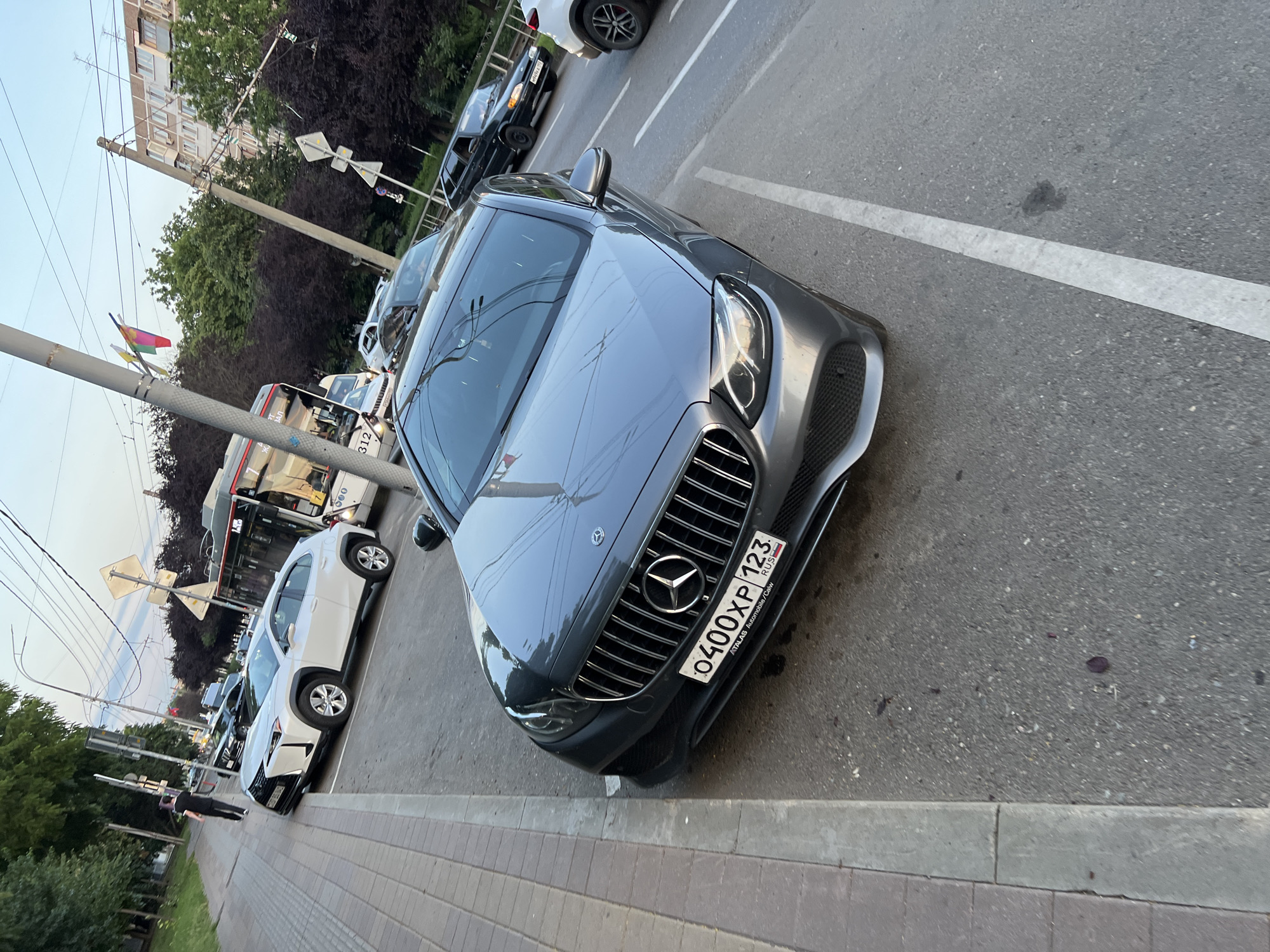 The image depicts a street scene in Krasnodar, Russia. In the foreground, a gray Mercedes-Benz E-Class sedan is parked, facing left. Its license plate reads "0400XP 123". To the left of the Mercedes, a white Lexus UX is parked. Further in the background, a bus, a black Lada car, and another white car are visible. A person is walking on the sidewalk on the far left of the image. The environment includes trees and buildings, typical of an urban setting. The sky suggests it is late afternoon or early evening.