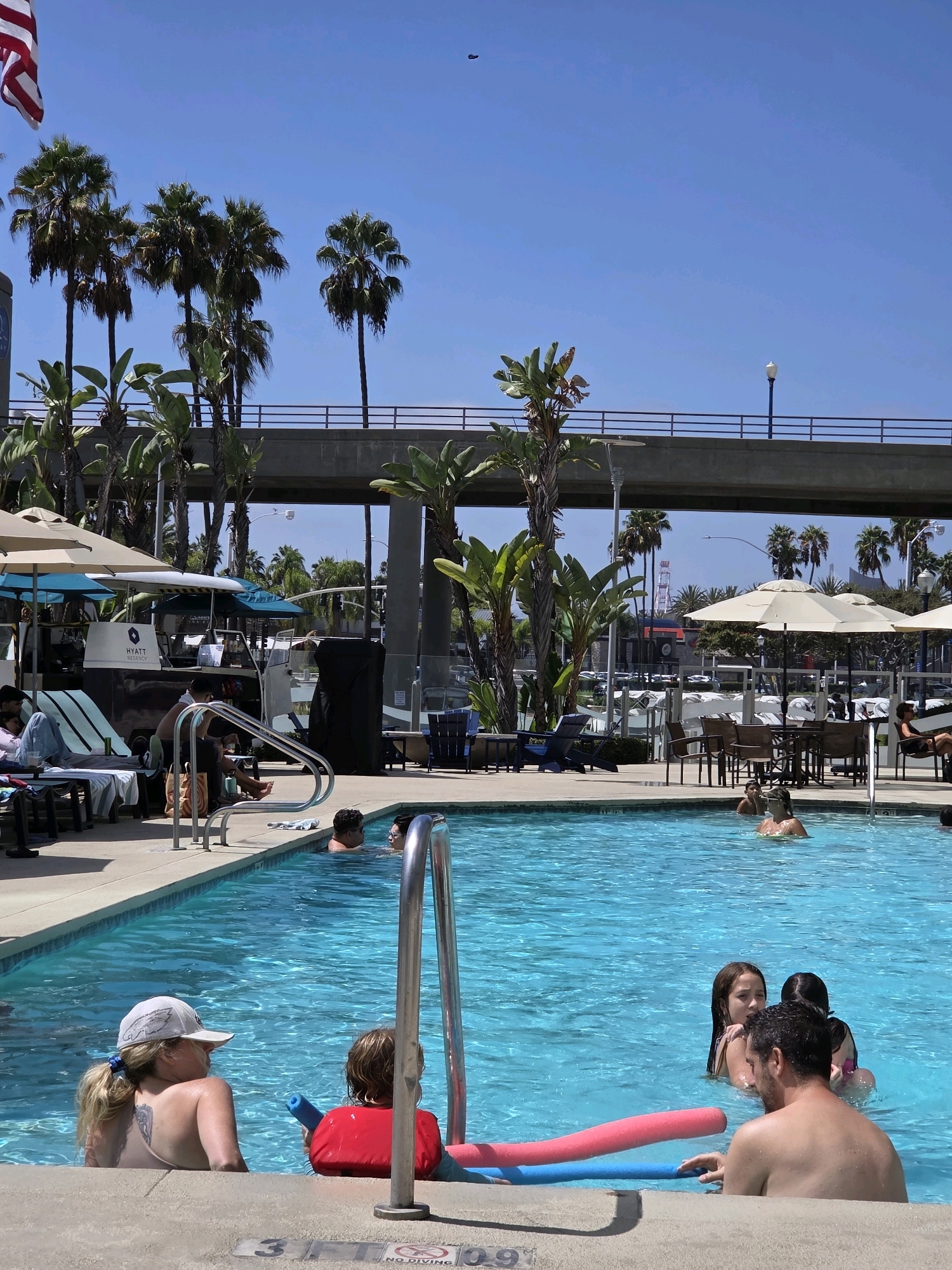 A group of people are enjoying a sunny day at a swimming pool located at the Hyatt Regency Long Beach, in Long Beach, California. The pool is a vibrant turquoise blue, with the edge of the pool and concrete decking visible in the foreground. In the pool, several individuals are present. Two young girls are swimming, with one wearing a pink swimsuit and the other a dark swimsuit. A man with a beard is also in the pool, near the girls. Closer to the edge, a woman with blonde hair, wearing a white baseball cap, is in the pool, facing away from the camera, with a young child in a red floatation device next to her. The child is holding a blue pool noodle. In the background, a Hyatt Regency shuttle is visible, along with palm trees, lounge chairs, and umbrellas. An overpass with a railing is in the upper background, with the bright blue sky above. An American flag is also visible in the upper left corner. The overall scene depicts a relaxed, recreational atmosphere typical of a hotel pool on a summer day.