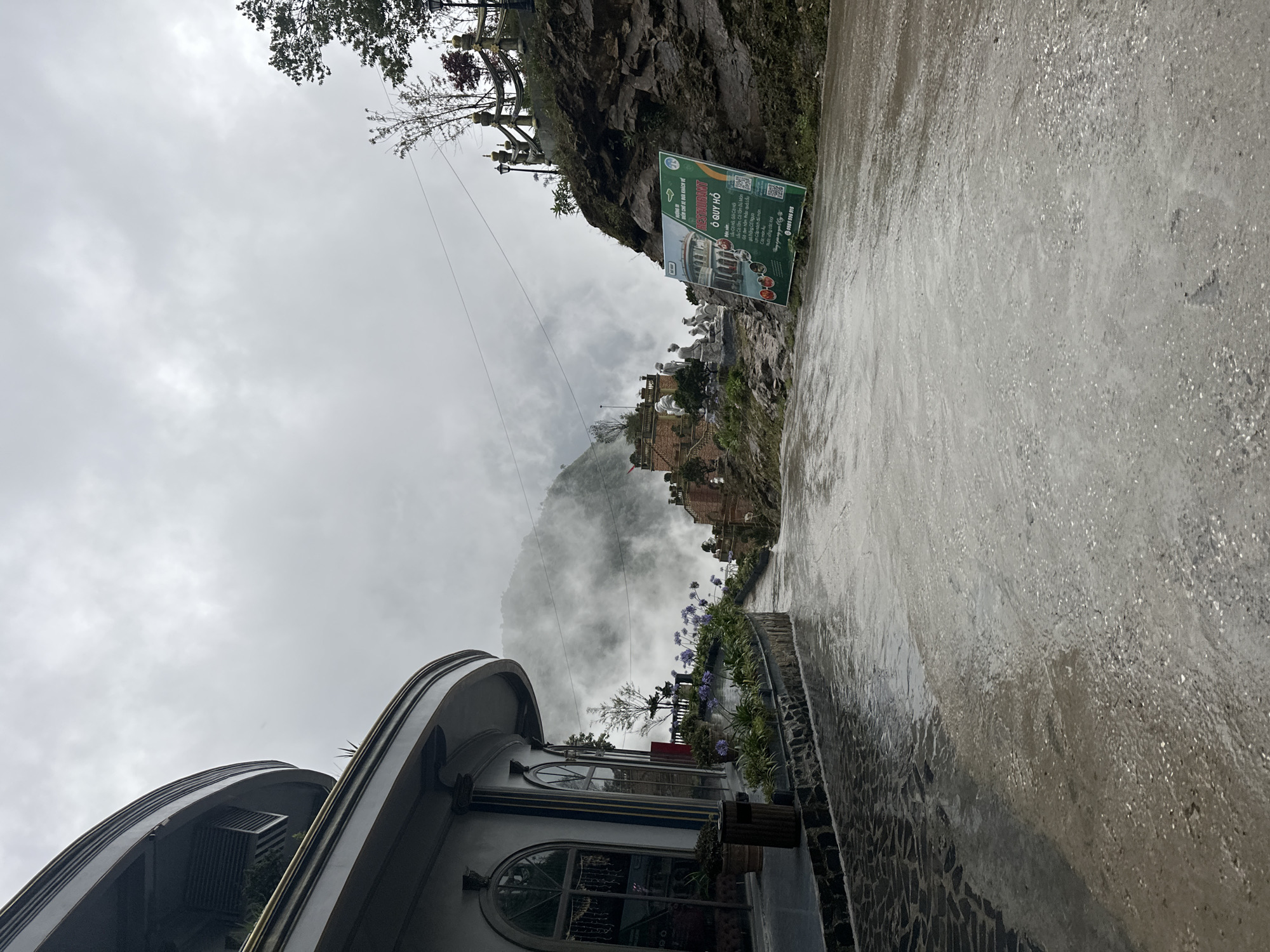 The image presents a slightly rotated view of a scene in Binh Lu, Vietnam. In the background, a misty mountain partially obscured by low-lying clouds dominates the view. Various buildings and structures are built into the side of the mountain. A sign advertising the "O Quy Ho Restaurant" is visible, along with details about the establishment. The foreground includes a building with a curved roof and ornate windows, possibly part of a restaurant or tourist establishment. A paved pathway is shown; the pavement is wet, likely due to rain. Purple flowers grow along the edge of the walkway. Power lines traverse the image horizontally. The sky is overcast, contributing to the overall cloudy atmosphere.