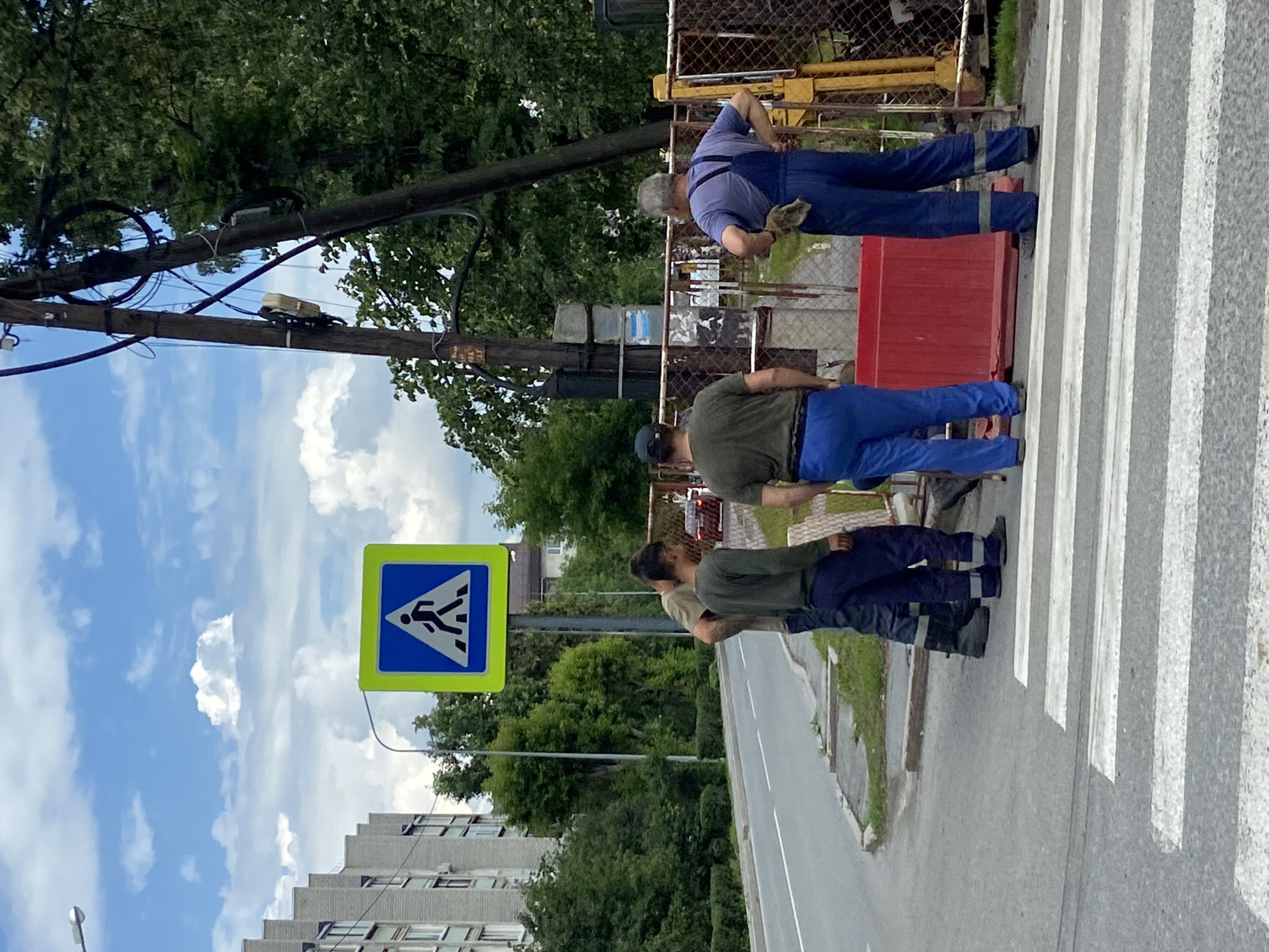 A group of four men in work clothes stand near a pedestrian crossing on a street in the city of Tyumen, Russia. One man, wearing a blue jumpsuit and a gray shirt, stands with his hands on his hips, facing away from the camera. Two other men, wearing dark green shirts and blue pants, stand facing the pedestrian crossing, one looking towards the road and the other looking down. A fourth man, wearing a dark green shirt and blue pants, stands slightly behind them, also facing the pedestrian crossing. A bright blue pedestrian crossing sign with a white pedestrian silhouette is visible behind the men. The street is paved and has a white line marking for the pedestrian crossing. In the background, there is a building with a grey facade, trees, a utility pole with wires, and a metal fence. A red portable barrier is also present on the sidewalk. The sky is partly cloudy.