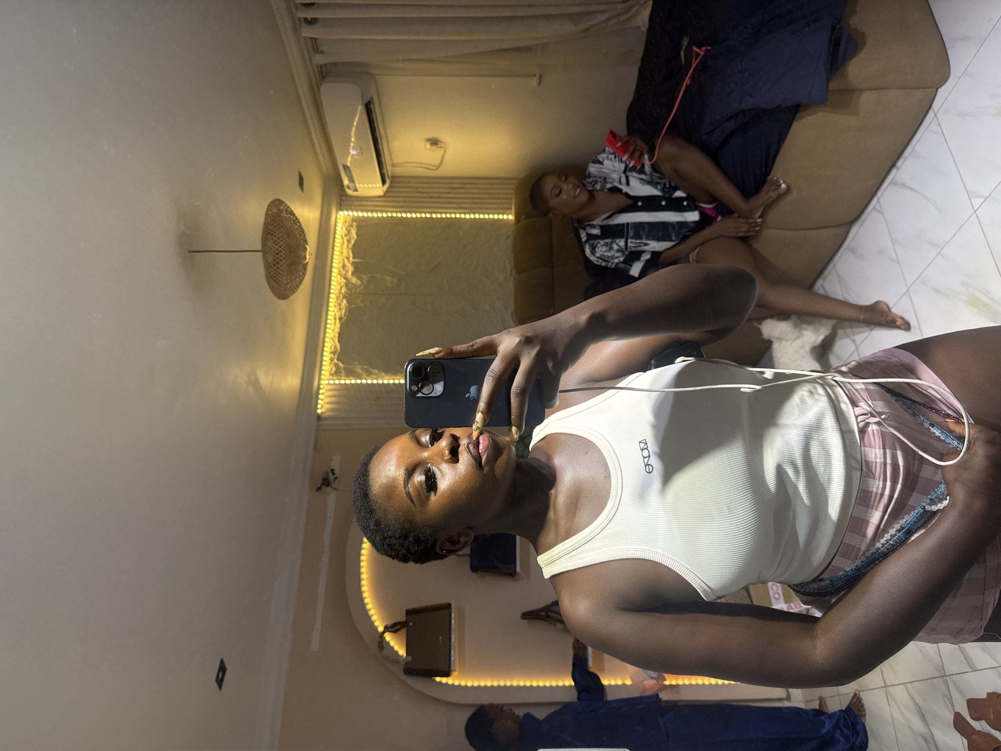 In a bedroom located in Abuja, Nigeria, a young woman is taking a selfie in a mirror. She is wearing a white tank top and pink plaid shorts. Her hair is short and dark, and she has on makeup. In the background, another woman is sitting on a bed, looking at her phone. The room is decorated with string lights, and there is an air conditioning unit on the wall. A handbag hangs on a shelf in the background, and a woven light fixture hangs from the ceiling. A white dog is partially visible in the lower left corner of the image.