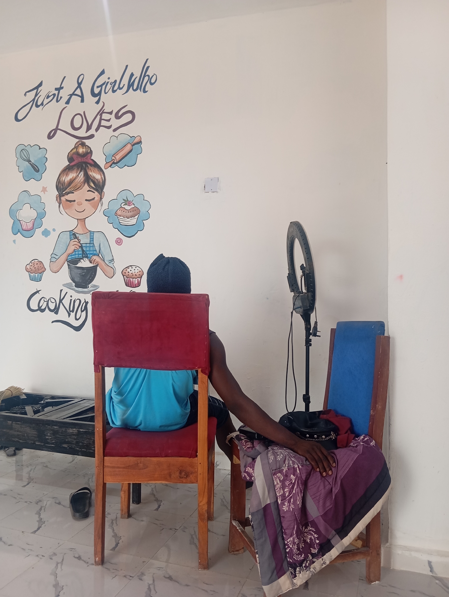 The image depicts an indoor scene in Gombe, Nigeria. A person wearing a blue shirt and a dark head covering is seated in a red cushioned chair. Next to them, another wooden chair with a blue cushion has a ring light and a purse on top of a purple and white patterned blanket. On the wall behind the chairs, there is a cartoon mural depicting a girl cooking with the text "Just A Girl Who LOVES Cooking" above it. A black sandal is on the floor to the left of the chair and a dark metal object is behind that. The floor is white with a marble pattern. The walls are white.