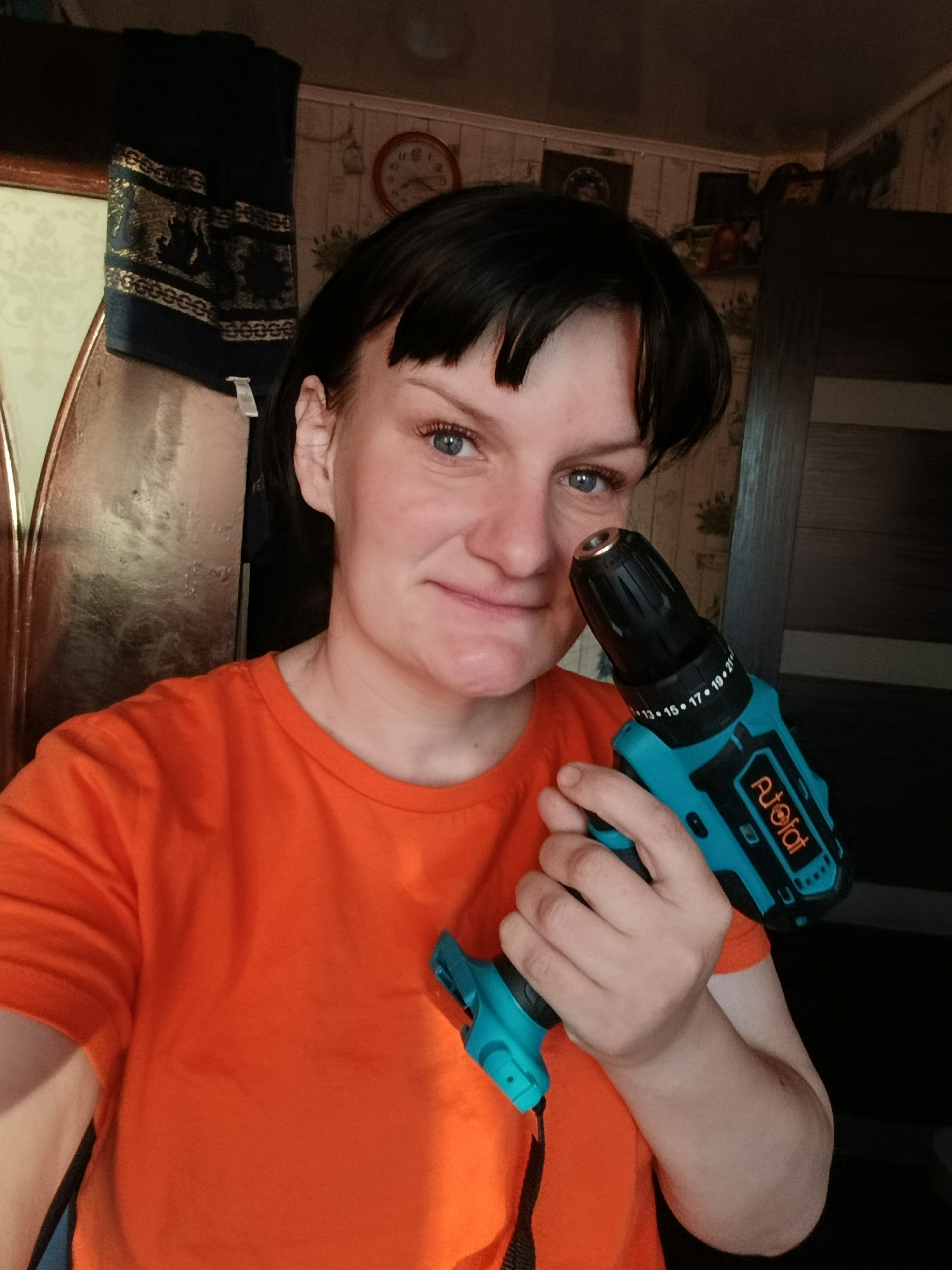 A woman with short, dark hair and bangs, and light blue eyes, is captured in a medium close-up selfie in an indoor setting. She is wearing a bright orange t-shirt and holds a teal and black cordless drill/driver in her right hand, angled slightly towards the camera. She has a subtle smirk or pleased expression on her face, with her eyes looking directly at the viewer.
The drill features the brand name "AUOIOF" prominently displayed in white text on its side. Around the chuck, numbers indicating settings or torque are visible, including "13, 15, 17, 19, 21".
The background suggests a domestic environment, possibly a home in Фоки, Russia. Light-colored, patterned wallpaper with what appears to be a birdcage design is visible. To the left, a dark blue fabric adorned with gold, intricate patterns hangs, possibly part of a curtain or furniture decoration. A round wall clock with red hands and numbers can be seen partially on the wall in the upper right quadrant. Dark wooden furniture, resembling a wardrobe or door, is also visible on the right side of the frame. The lighting indicates it is likely daytime indoors.