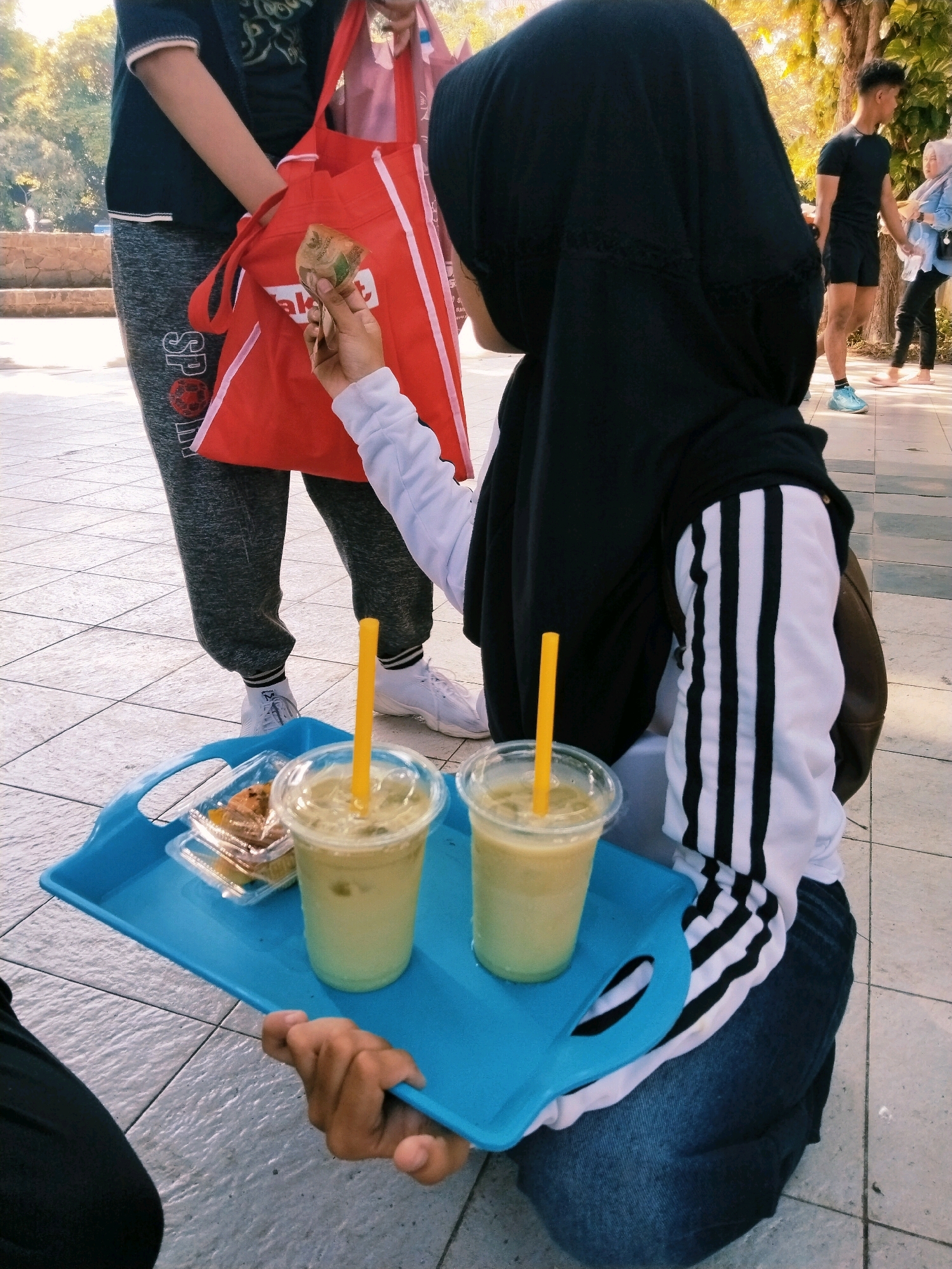 A person wearing a black hijab and a white long-sleeved shirt with black stripes is holding a blue tray with two drinks and a small container of food. The person is kneeling on a tiled surface. In the background, another person wearing a black t-shirt and shorts is standing, and to their right, another person is walking. The environment appears to be an outdoor urban setting, possibly a public space or park, in Surabaya, Indonesia, suggested by the presence of trees and tiled paving.