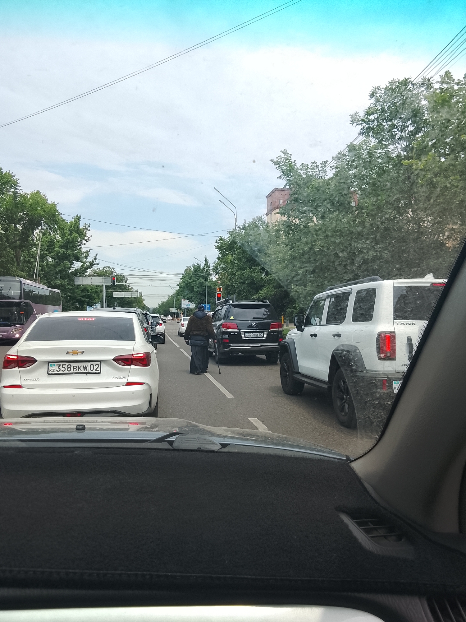 The image depicts a congested street scene in Almaty, Kazakhstan, characterized by heavy traffic. The perspective is from inside a vehicle, looking forward through the windshield. In the foreground, the dashboard and windshield wipers of the vehicle are visible.
The roadway is lined with multiple cars in stationary traffic. A white Chevrolet sedan with a license plate reading "KZ 358BKW 02" is prominent in the center-left of the frame. To its right, a white SUV, identified by the name "TANK" on its rear, is also in traffic. Further down the road, a black Lexus SUV is visible.
A lone figure, an elderly person wearing dark, flowing clothing and a headscarf, is walking across the street, utilizing a cane for support. They are positioned between lanes of traffic, appearing to navigate the congestion. Traffic lights are visible in the distance, indicating an intersection.
The environment is urban, with trees lining the sides of the street and buildings visible in the background. The sky is overcast with a mix of blue and white clouds. The overall impression is that of a typical city traffic jam under cloudy weather conditions in Almaty.