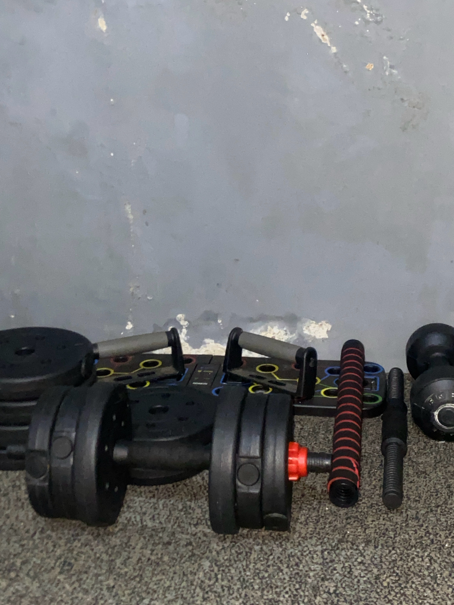 This image depicts a collection of weightlifting equipment arranged on a carpeted floor against a textured, gray wall. The equipment includes a pair of adjustable dumbbells, a push-up board with various colored grip positions, and weight plates. The scene is static and appears to be an indoor setting, possibly a home gym or a fitness studio. There are no people or visible actions taking place. The location is identified as Batam, Indonesia.