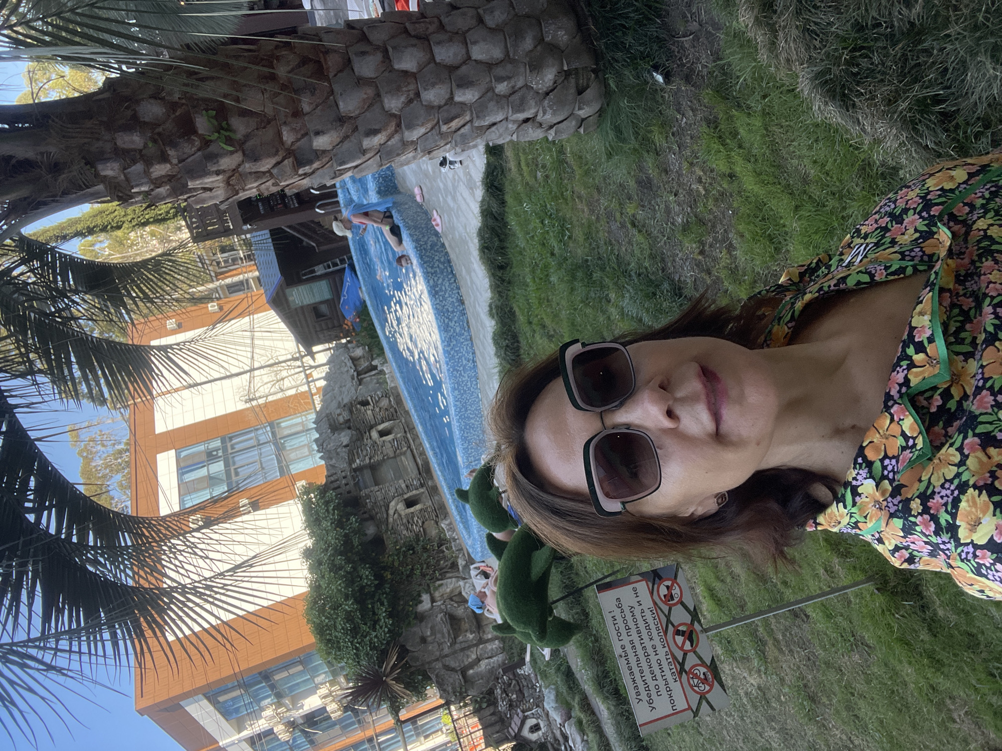 A medium shot captures a woman with brown hair and sunglasses taking a selfie in front of a swimming pool and palm trees. She is wearing a floral print shirt with green trim. In the background, a modern building with orange and white facades and blue windows stands behind a lush green area with decorative topiaries shaped like dolphins. A small wooden cabin and a stone-like structure are also visible near the pool. A sign with Russian text and pictograms indicating no bicycles, skateboards, or strollers is positioned in front of the woman. Two people are in the pool, one of whom appears to be a child. The setting suggests a recreational area, possibly a hotel or resort in Sochi, Russia, given the presence of palm trees and the architectural style.