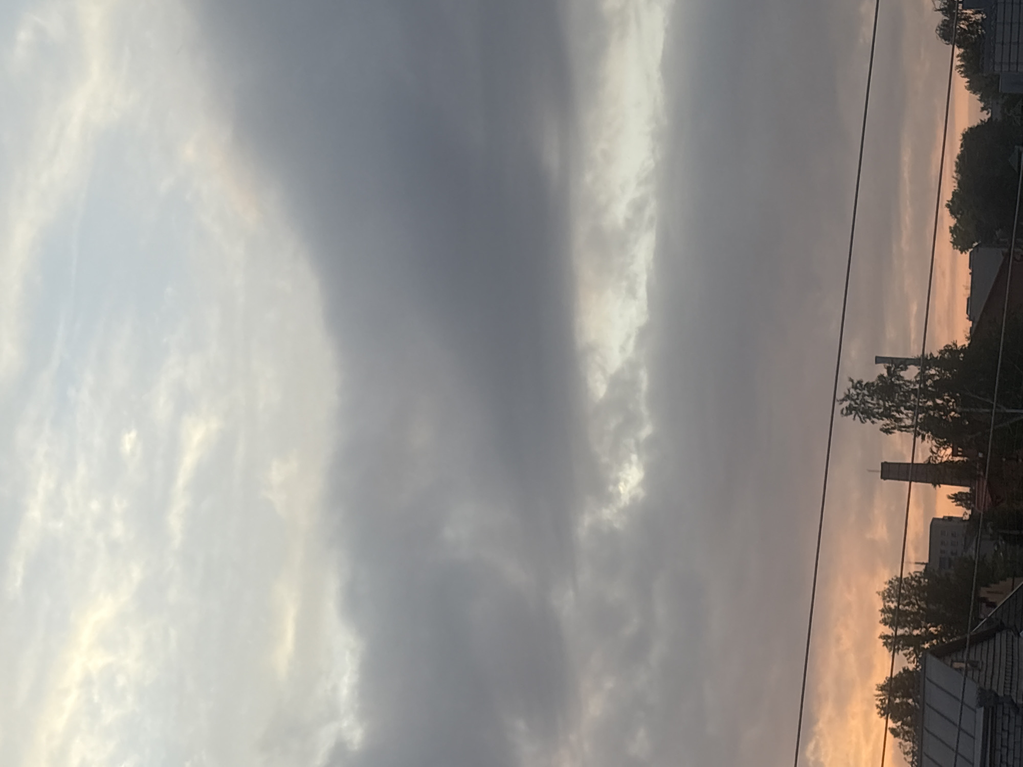 The image shows a cloudy sky with a hint of sunset colors on the horizon. In the foreground, there are power lines stretched across the frame, and below them, the rooftops of buildings and trees are visible. One building appears to have a tall, industrial chimney. The sky is filled with varying shades of grey and white clouds, with some lighter areas suggesting the sun is low in the sky. There are no people or animals visible in the image. The scene appears to be a view of an urban or suburban landscape at dusk.