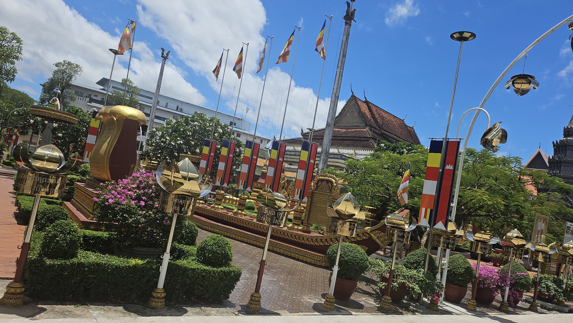 This image captures the vibrant grounds of Wat Preah Prom Rath in Siem Reap, Cambodia, on a sunny day. The foreground is dominated by decorative golden structures resembling lotus flowers atop poles, with intricate carvings in Khmer script. Flanking these are neatly trimmed green bushes and potted plants filled with bright pink flowers.
Behind the decorative elements, a large, ornate golden boat-like structure rests on a pedestal, adorned with colorful Buddhist flags. The flags, featuring horizontal bands of yellow, red, blue, and white, are a common sight at Buddhist temples. These flags are also seen flying from tall poles scattered throughout the scene, interspersed with solar-powered lamps and artistic metal sculptures that resemble birds.
In the mid-ground, a traditional Cambodian temple building with a red tiled roof and intricate architectural details stands prominently. Adjacent to it, a modern, multi-story white building with prominent blue accents is visible. Lush green trees with white blossoms frame the temple and the modern structure, creating a beautiful contrast.
The sky above is a clear, bright blue, dotted with fluffy white clouds, indicating fair weather. The ground appears to be paved with reddish-brown bricks and concrete in various sections. The overall scene is one of religious and cultural significance, showcasing the blend of tradition and modernity within the temple complex. No people are visible in the frame.