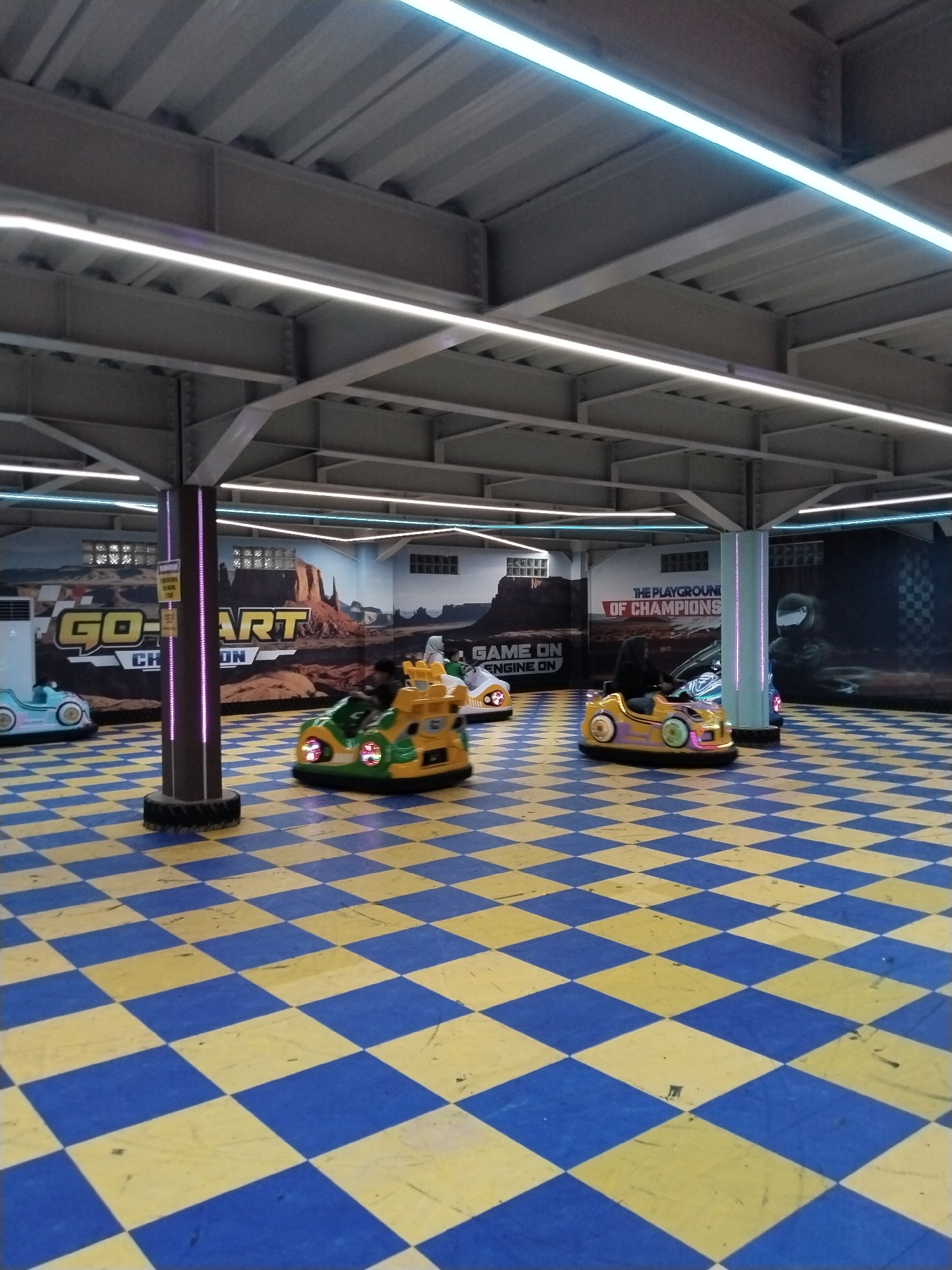 The image depicts an indoor go-kart arena with a checkered yellow and blue floor. Several bumper cars are visible, with two in motion. One bumper car, colored green and yellow, has a person inside. Another, predominantly yellow with purple accents, is also occupied. In the background, there are murals depicting a desert landscape and the words "GO-KART CHAMPION" and "THE PLAYGROUND OF CHAMPIONS." The arena is illuminated by fluorescent lights lining the ceiling structure. The scene appears to be located in an entertainment venue.