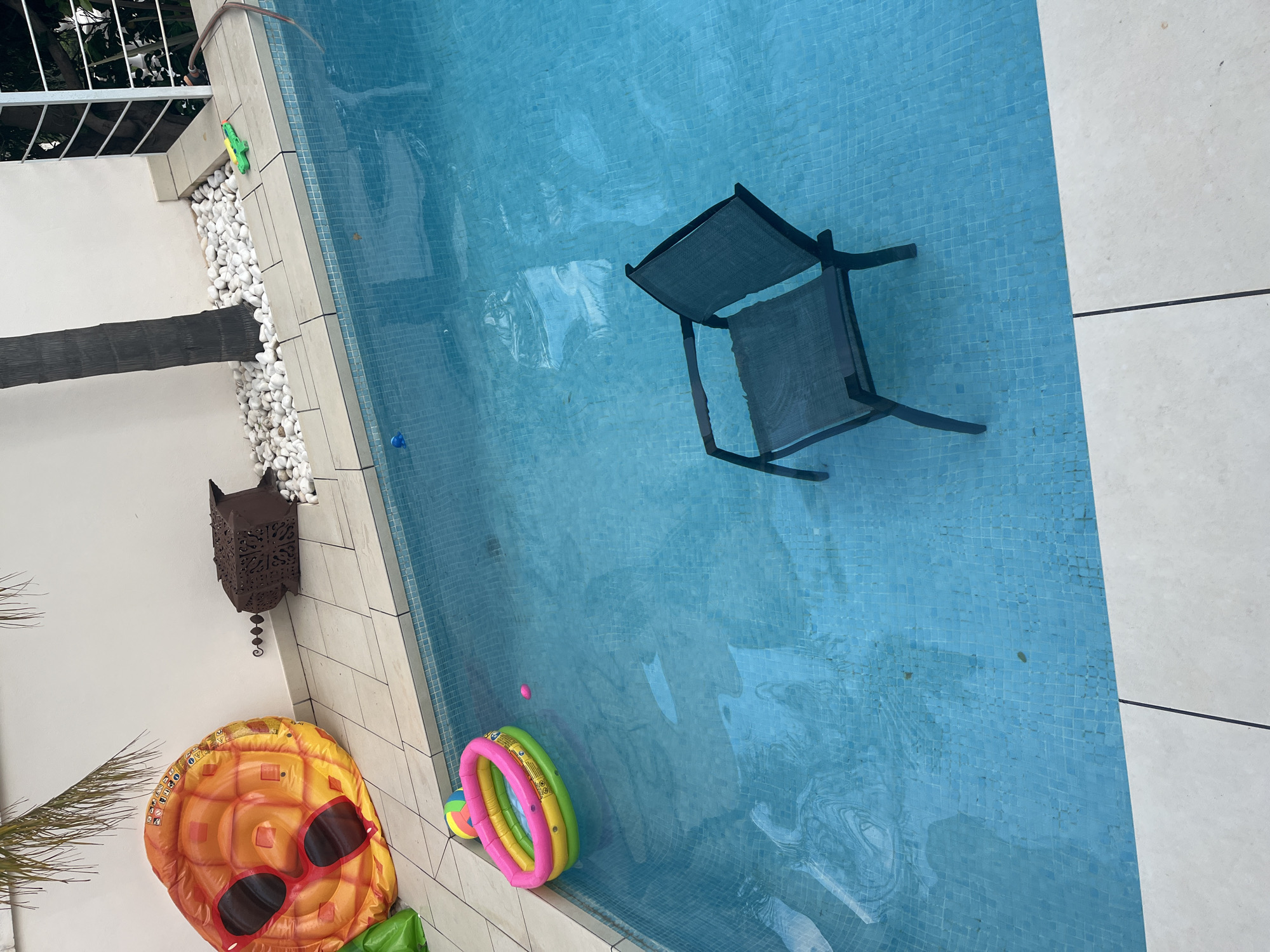 The image shows a swimming pool in El Poble Nou de Benitatxell / Benitachell, Spain. The pool water is light blue and reflects the sunlight. A black folding chair is submerged in the water. On the pool deck, there's a pineapple-shaped inflatable pool float with sunglasses, a colorful ring-shaped inflatable toy, and a lantern hanging on the wall. White decorative pebbles line the edge of the pool. A green inflatable crocodile is also visible. The pool is surrounded by white tiled surfaces. A black palm trunk is located in the corner of the pool area. A section of a white metal railing is partially visible at the top of the frame.