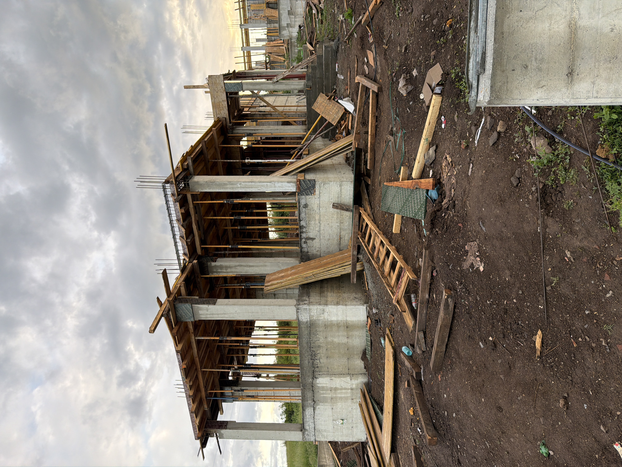 The image shows a building under construction in what appears to be a rural area. The building is a two-story structure with concrete pillars and wooden framing. Rebar extends from the top of the structure. Scaffolding and other construction materials are scattered around the site. There are some other building under construction in the distance. The location is unidentified.