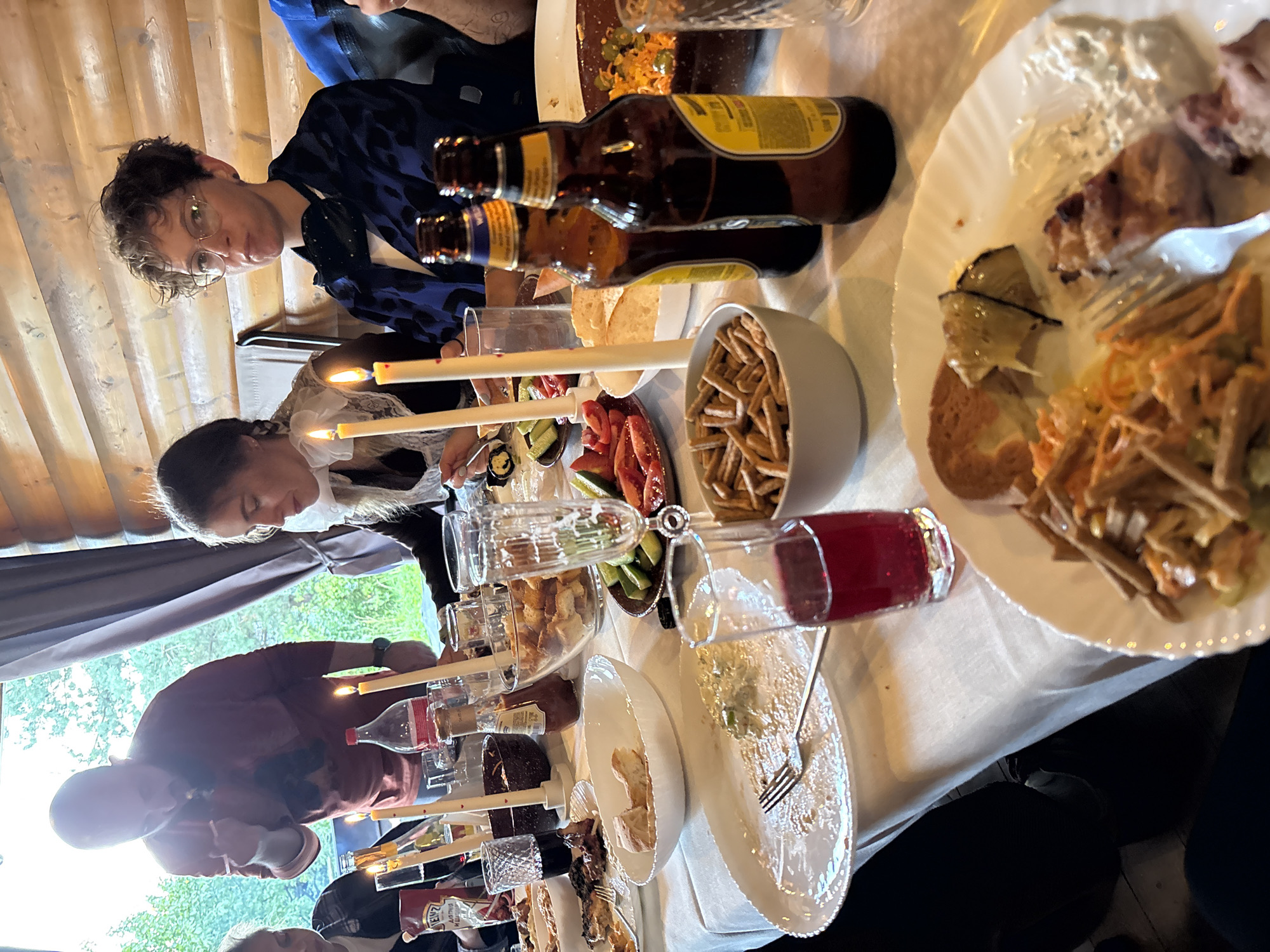 The image depicts a group of people seated at a table laden with food and drinks, possibly in a rustic wooden cabin. The setting appears to be Levashovo, Russia, given the context. A man in a reddish-brown shirt stands and leans over the table, seemingly engaged in conversation or serving food. To his right, a woman with a white ruffled collar looks down at her plate. Across the table, a person with curly hair and glasses is looking forward with their mouth slightly open. The table is covered with a white tablecloth and features several plates of food, including what appears to be meat, vegetables, and bread. There are also bottles of beer, glasses of red liquid, and lit candles. The background shows a wooden wall and a window looking out onto greenery. The overall scene suggests a casual gathering or meal.