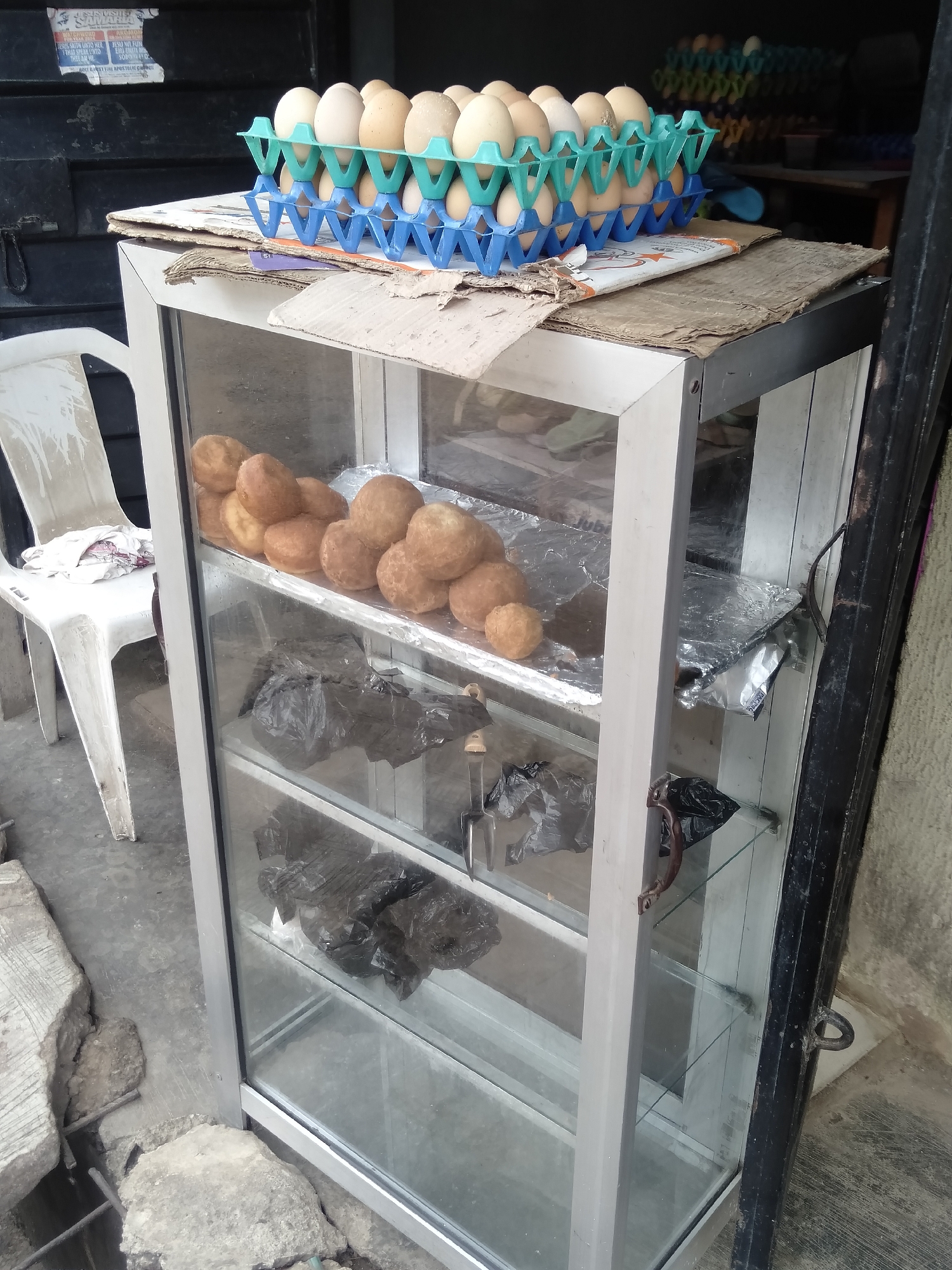 This image captures a street vendor's display cabinet in Shangisha, Nigeria. The central focus is a silver-framed glass display case, likely used for food sales.
On top of the cabinet, stacked on corrugated cardboard, are two plastic egg trays, one blue and one teal, filled with light brown eggs. More egg trays are visible in the blurred background.
Inside the top shelf of the cabinet, a pile of round, light-brown fried dough balls (likely 'puff-puff' or 'akara') is displayed on aluminum foil. Below this, on the middle shelf, are several items wrapped in black plastic bags and aluminum foil. The bottom shelf is empty. Visible within the cabinet on the right side are kitchen tools, including tongs or a spatula.
To the left of the cabinet, a white plastic chair rests on a concrete ground with exposed rebar and broken concrete pieces. A white cloth or towel is draped over the chair. A black metal frame or door is partially visible on the right side of the image.
In the background, behind the eggs, a faded and torn poster is affixed to a dark surface. Visible text on the poster includes: "JESUS SAITH UNTO HER", "THAT SPEAK INTO", "JESU WEFUN", "EMI NITI MO", "SOROO", and "HOLY GHOST FIRE APOSTOLIC CHURCH". The word "FACPRO" is also faintly visible on a piece of cardboard on top of the cabinet.
The scene suggests a daytime setting for an informal food stall or market stand.