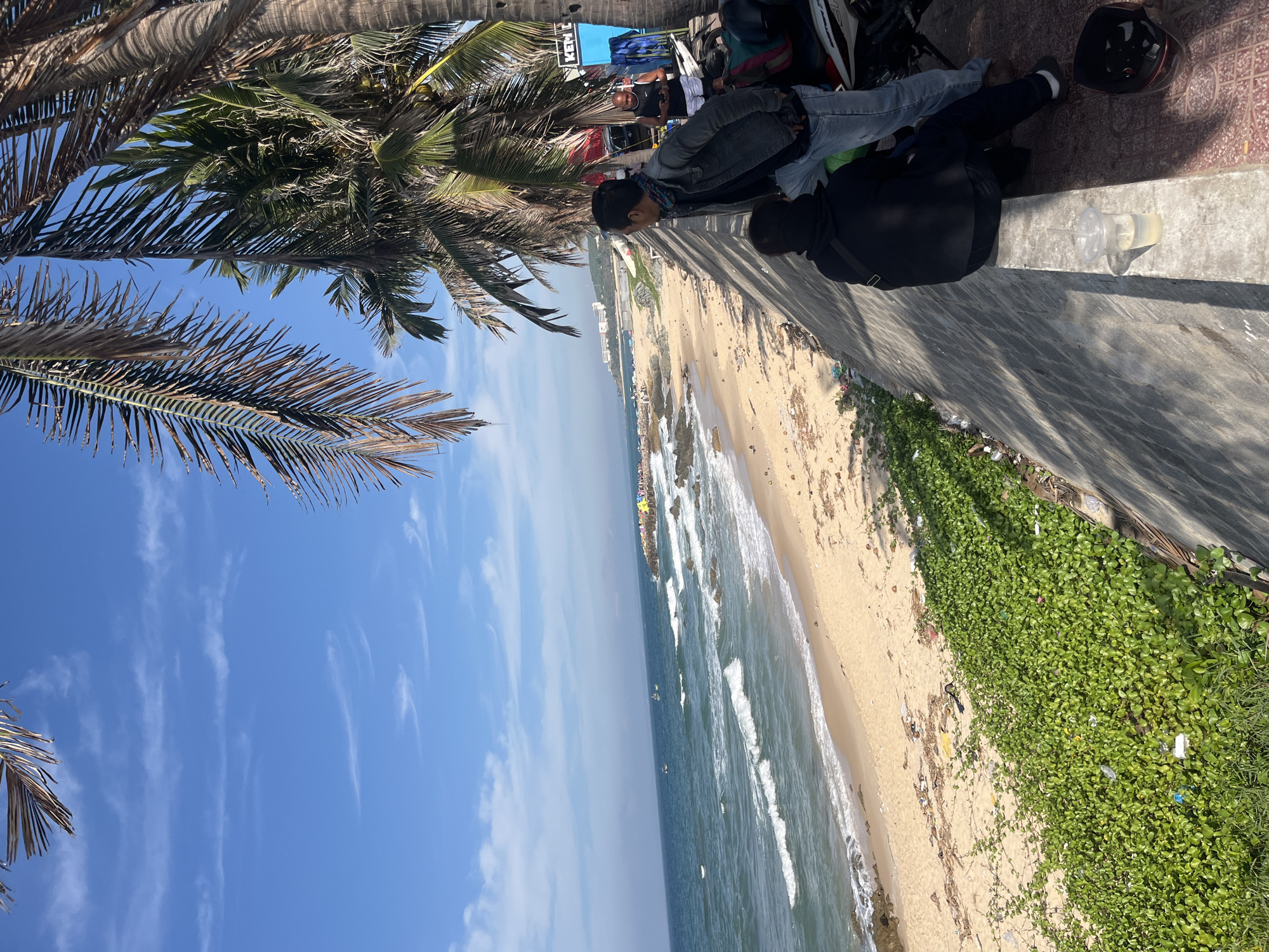 The image depicts a scenic view of a beach in Phu Thuy Ward, Vietnam. The foreground features a low concrete wall with lush green vegetation growing along its base, interspersed with some scattered debris. Two individuals are present, with one seated on the wall and the other standing beside them, both facing towards the ocean. They appear to be observing the seascape. Further back, a sandy beach stretches along the coastline, with gentle waves lapping at the shore. Several small boats are visible in the water, and a rocky pier with colorful flags extends into the sea. Palm trees with fronds frame the top of the image, suggesting a tropical or subtropical climate. In the background, buildings can be seen on a distant hillside. A parked motorcycle and a helmet are also present near the standing individual, indicating a mode of transportation. The sky is blue with scattered white clouds, indicative of a clear day.