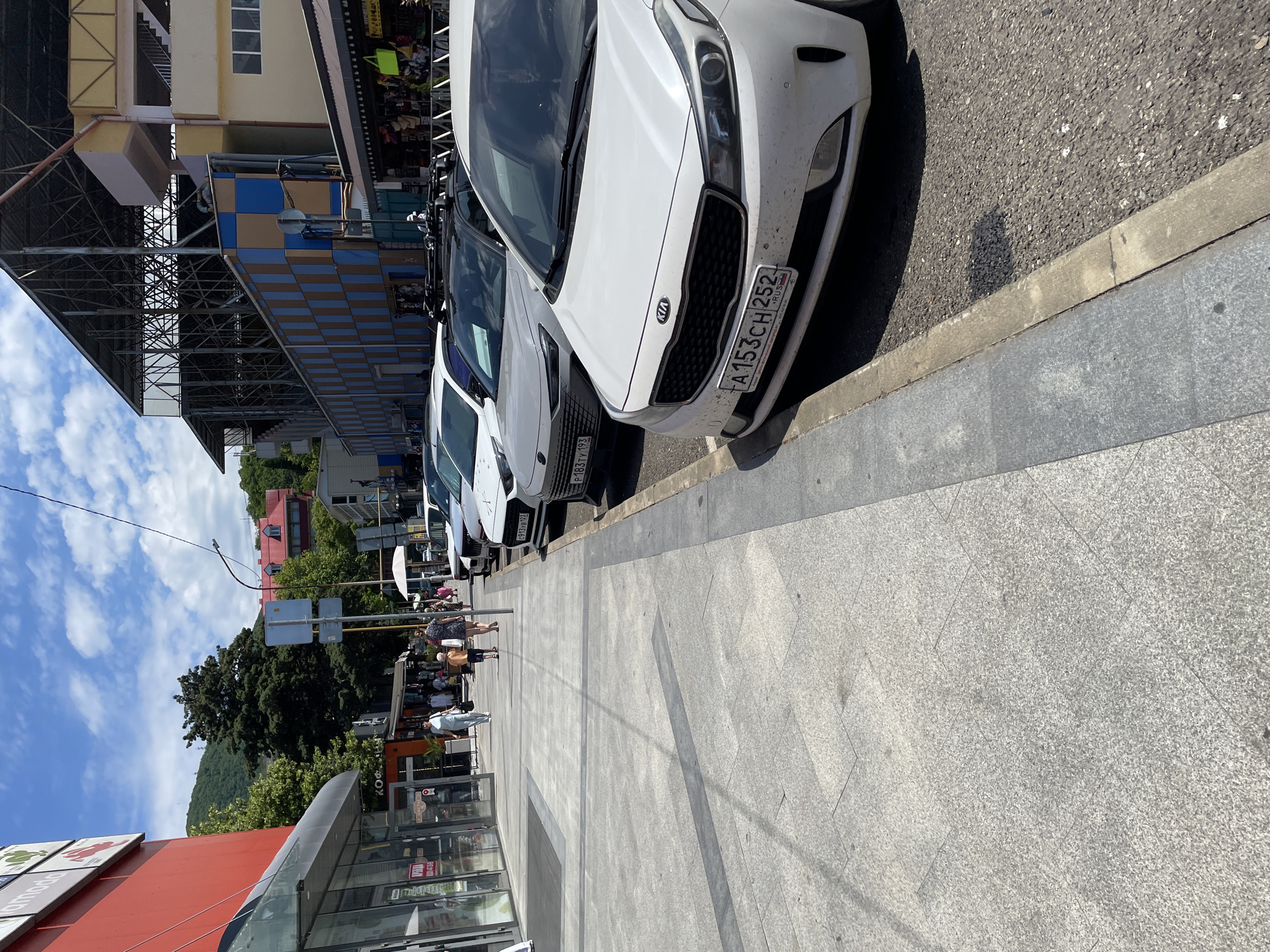 The image depicts a street scene on a bright, sunny day, likely in the afternoon given the strong shadows. The perspective is angled, looking down a sidewalk lined with parked cars. In the foreground, a grey tiled sidewalk leads towards a building with a red facade and large glass windows, one of which has some text visible, including "lamoda".
Several cars are parked along the curb, with a white Kia sedan prominently displayed. Its license plate is clearly visible: "A153CH 252 RUS". Behind the Kia, other cars are partially visible, including a lighter colored one with a license plate that appears to start with "P 183".
On the sidewalk, several people are walking and browsing stalls set up in front of shops. The people are dressed in casual summer clothing. One woman is wearing a dark patterned dress and carrying a bag, while another person is in an orange top and shorts. Further down, a man in a light blue shirt is walking towards the viewer.
The background shows a street with more shops and buildings, some with colorful checkered facades. Lush green trees and a distant, tree-covered mountain range are visible under a blue sky dotted with white clouds, suggesting a pleasant, warm climate. The overall scene conveys a bustling but relaxed atmosphere of a commercial area on a summer day.