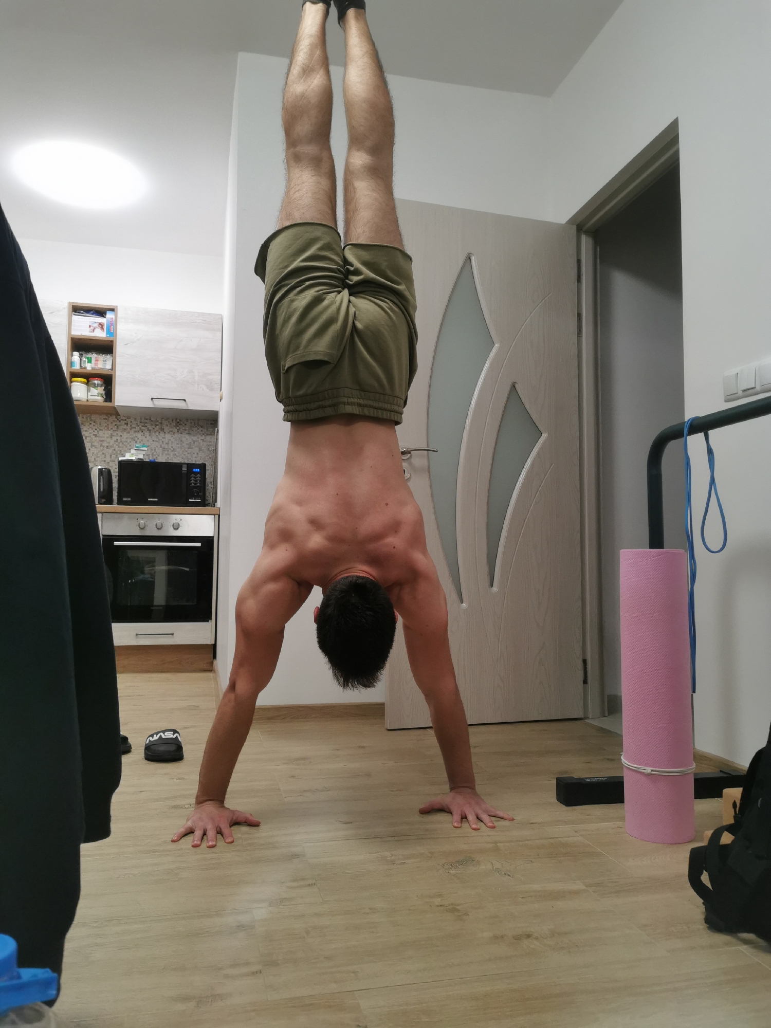 A person is performing a handstand in an indoor environment. The person is male, shirtless, wearing green shorts, and has dark hair. He is positioned upside down with his hands on a light-colored wooden floor.
Behind him, a partially visible kitchen area includes an oven, a microwave, and cabinets. A door with a decorative glass panel is visible to the right of the person. A pink yoga mat and a black pull-up bar are located near the door. A partially obscured black jacket and blue container are in the foreground. A pair of black sandals are located next to the jacket.
The location of the image cannot be determined based on the visual data available.