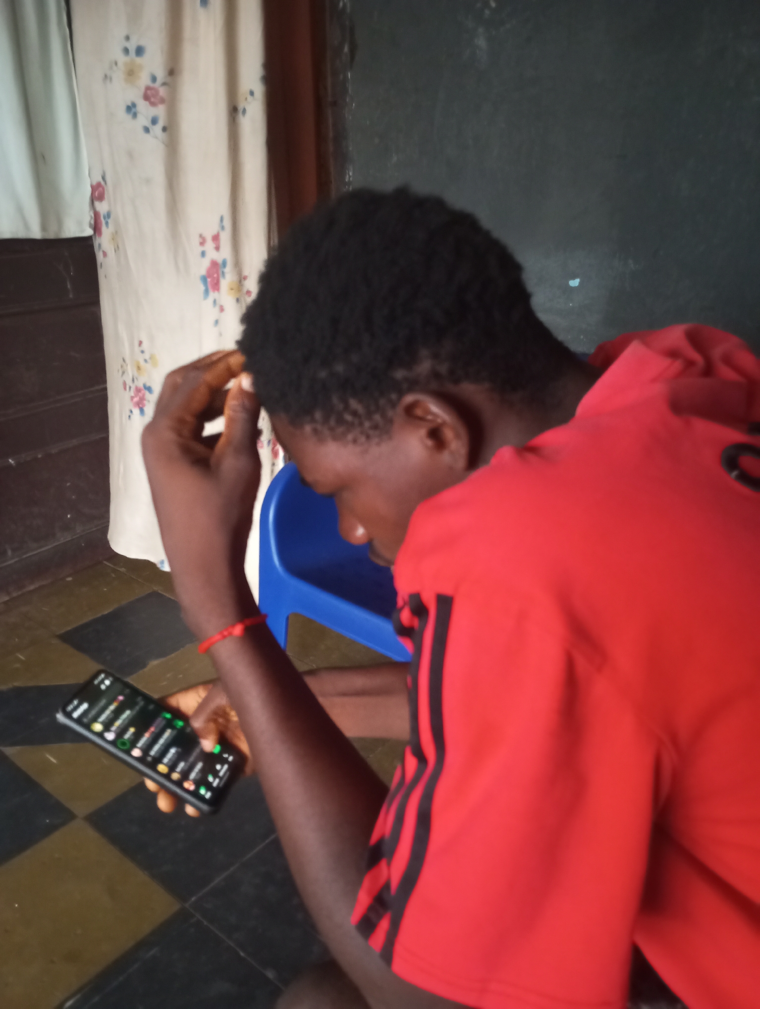 A young Black person is sitting on a blue plastic chair in an indoor environment. The person is wearing a red hooded sweatshirt with black stripes on the sleeves. They are holding a smartphone in their left hand, which displays a green-tinted user interface with multiple lines of text and icons. The person's right hand is touching their head, with their fingers partially in their hair, suggesting contemplation or concern. The background includes a beige curtain with a floral pattern, a brown door, and a black and yellow checkered floor. The lighting is dim, casting shadows and highlighting the subjects in the frame. The overall setting appears to be domestic.