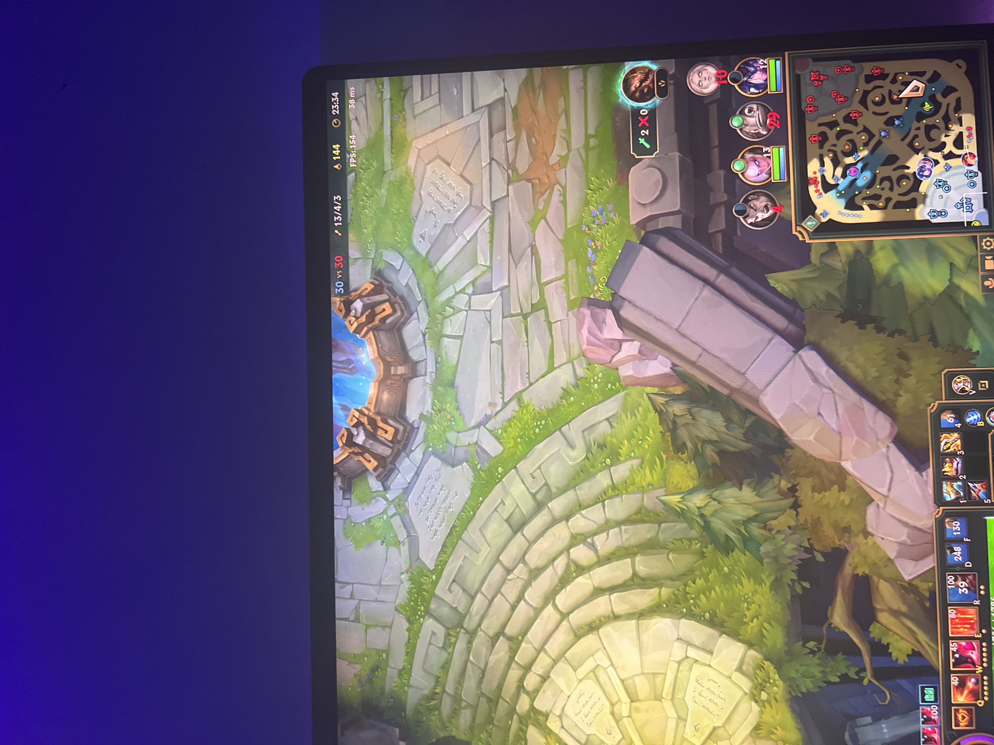 The image is a screenshot of a League of Legends match displayed on a monitor. The game is set in a fantasy environment with stone structures, grass, and trees. The top of the screen shows game statistics including the score (30 vs 30), player kill/death/assist count (13/4/3), gold (144), time (23:34), FPS (154), and ping (38 ms). The bottom right corner displays a minimap of the game world, with various icons indicating player positions and objectives. The bottom of the screen shows player ability icons and health bars. There are no people present in the image, nor any specific location within a city.