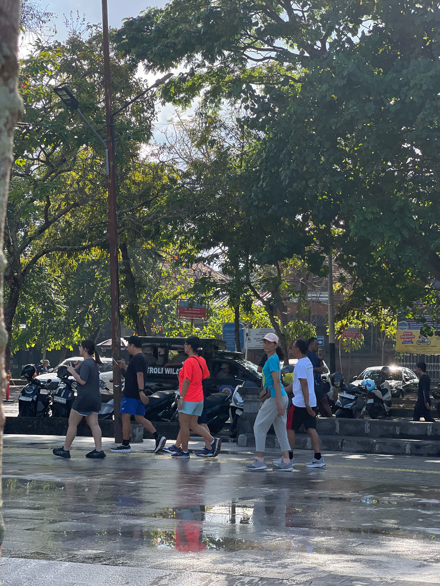In Denpasar, Indonesia, a group of six individuals, some appearing to be locals and others tourists, walk across a wet, reflective street. The group is composed of three women and three men, each dressed in casual, athletic-style clothing. The street is lined with parked motorcycles and cars, and the scene is framed by lush, green trees. Sunlight filters through the leaves, casting dappled shadows on the ground. The wet surface suggests recent rain or a street cleaning. The overall atmosphere is that of a bright, active day in a tropical city.