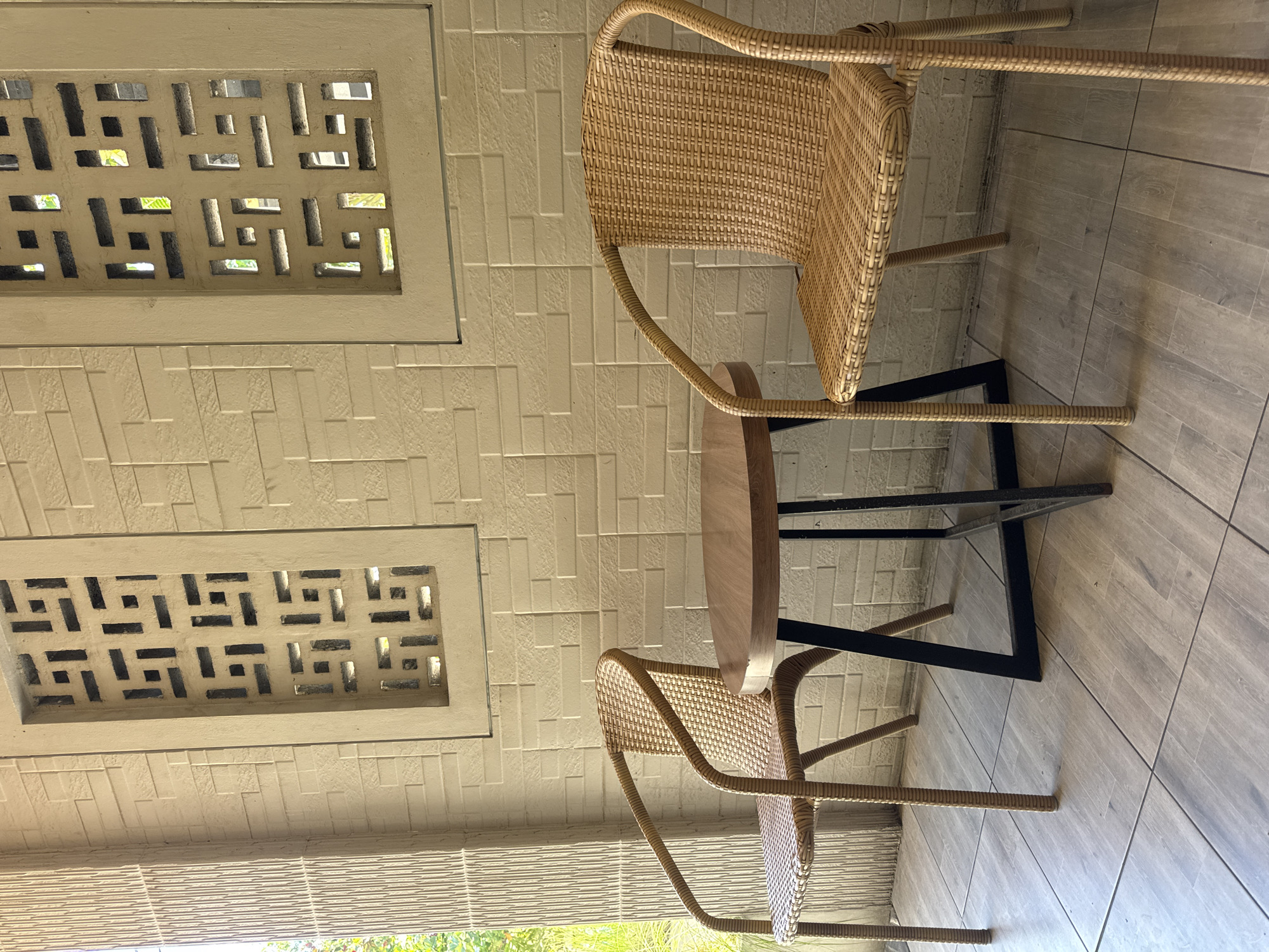 The image shows a small round wooden table with black metal legs positioned between two woven wicker chairs. The scene appears to be indoors, near a wall featuring two decorative rectangular ventilation blocks. The floor is tiled with wood-look planks. Based on the presence of outdoor wicker chairs and general architectural style, it is probable the location is Kuta, Indonesia.