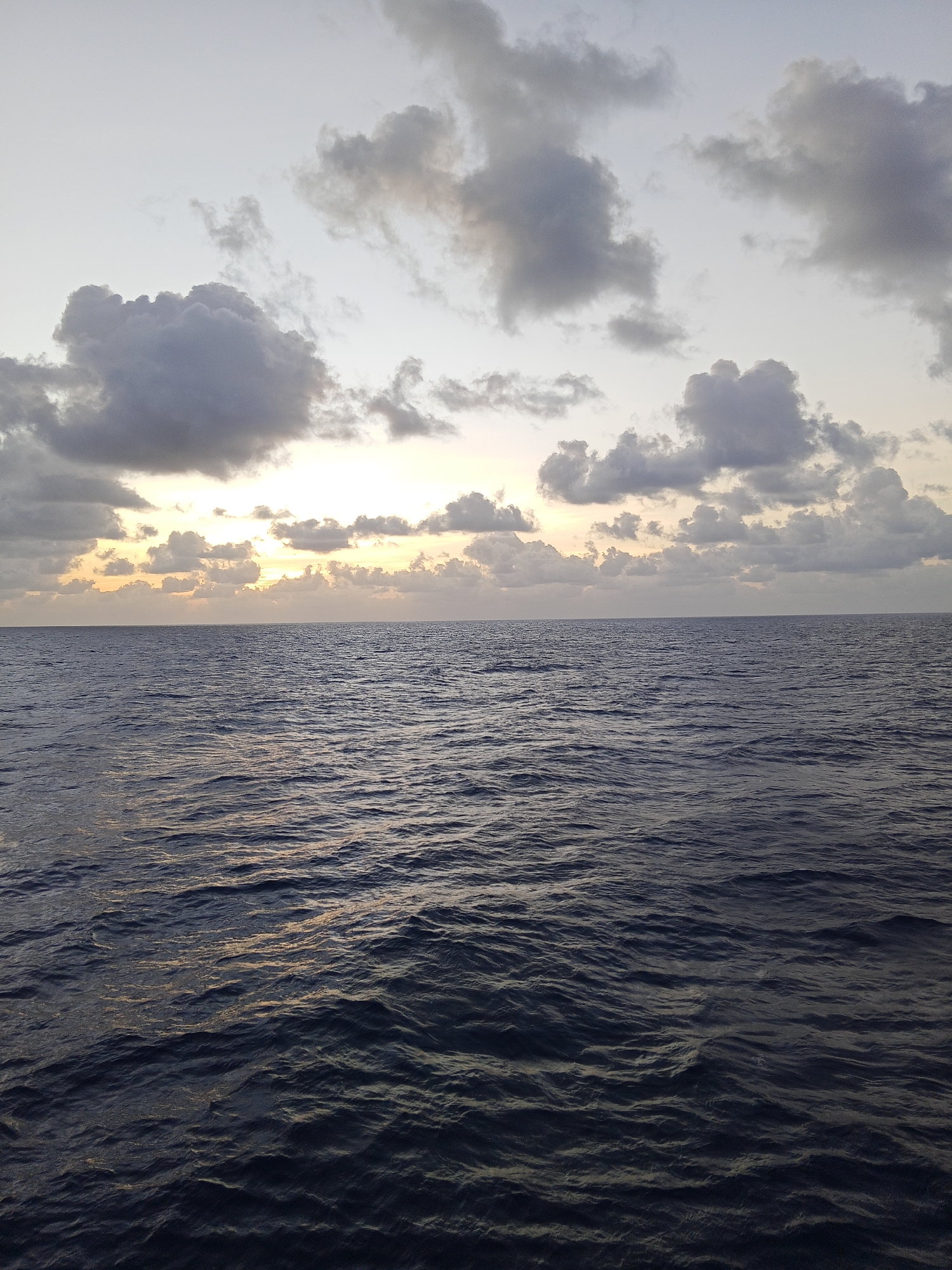 The image depicts a broad expanse of dark blue, choppy ocean water under a sky filled with scattered grey and white clouds. The sun is low on the horizon, creating a band of soft yellow and orange light peeking through the clouds. The lighting suggests either sunrise or sunset. The water is a deep, rich blue with visible ripples and small waves. The sky is a pale grey at the top, transitioning to lighter shades near the horizon where the sun is visible. The image is a landscape orientation and appears to be taken from a boat or similar vantage point on the water. There are no people or man-made structures visible in the frame.