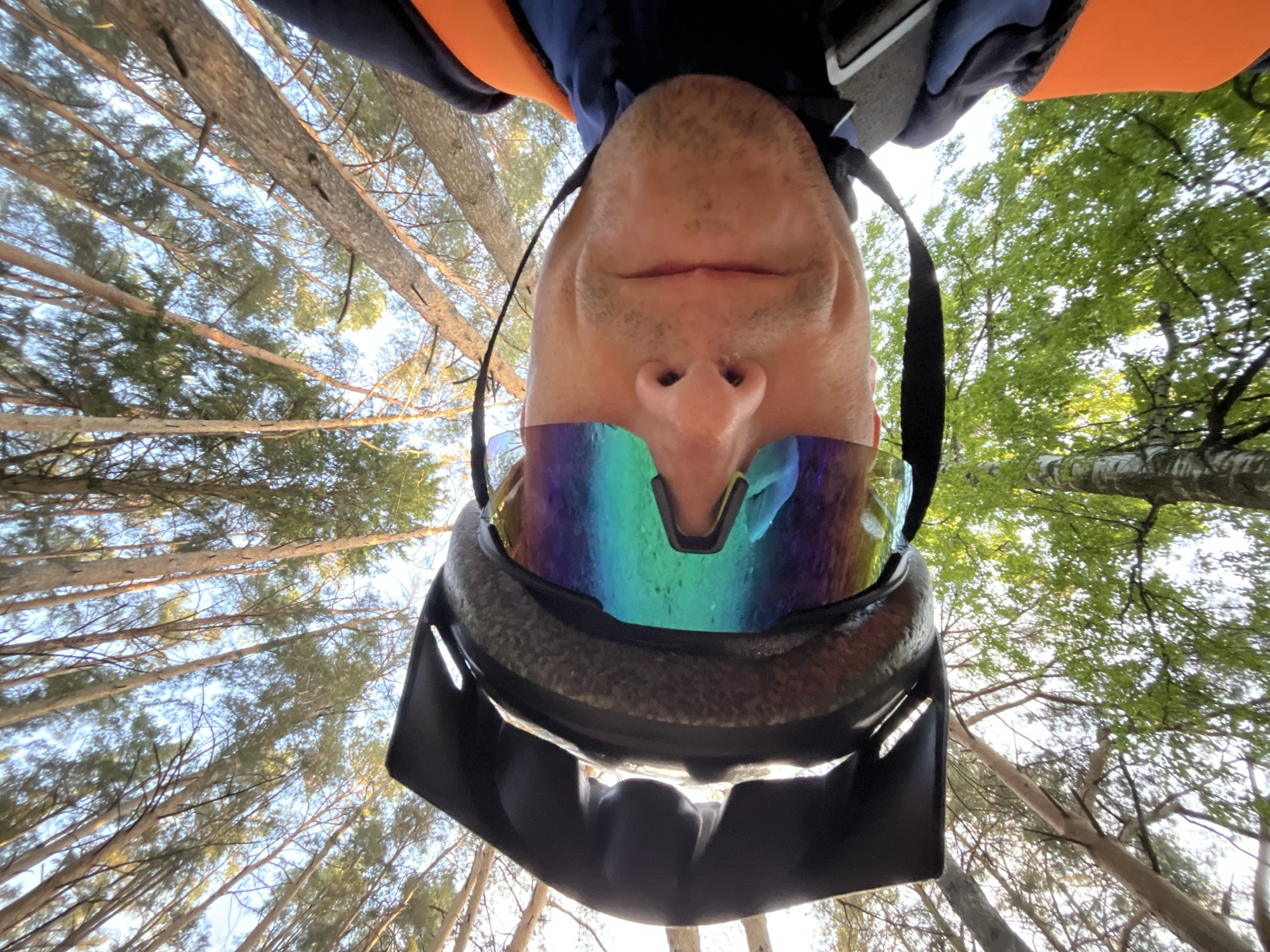 A low-angle perspective image shows a man wearing a helmet and iridescent goggles, positioned upside down relative to the surrounding environment. He is looking directly at the camera. The background consists of tall trees reaching upward, suggesting a forest setting. On the left side of the image, the trees appear thinner and more sparse, allowing glimpses of the sky. On the right side, the trees are denser with green foliage. The man is wearing an orange and blue athletic garment. The location is unclear, but this is unrelated to the city of Chaikovsky, Russia.