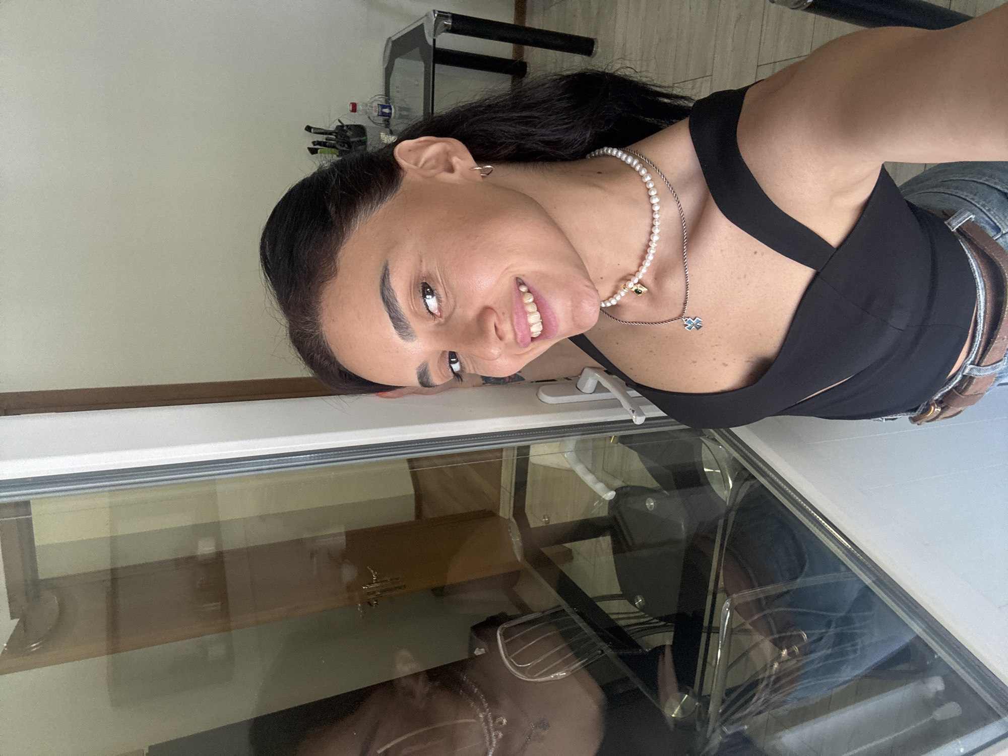 A young woman with dark hair and a bright smile takes a selfie in front of a glass door. She is wearing a black top, a pearl necklace, and hoop earrings. The reflection in the glass shows a room with a table and chairs. The scene appears to be indoors, possibly in an apartment or office. The image was captured at a close-up angle, focusing on the woman's face and upper body.