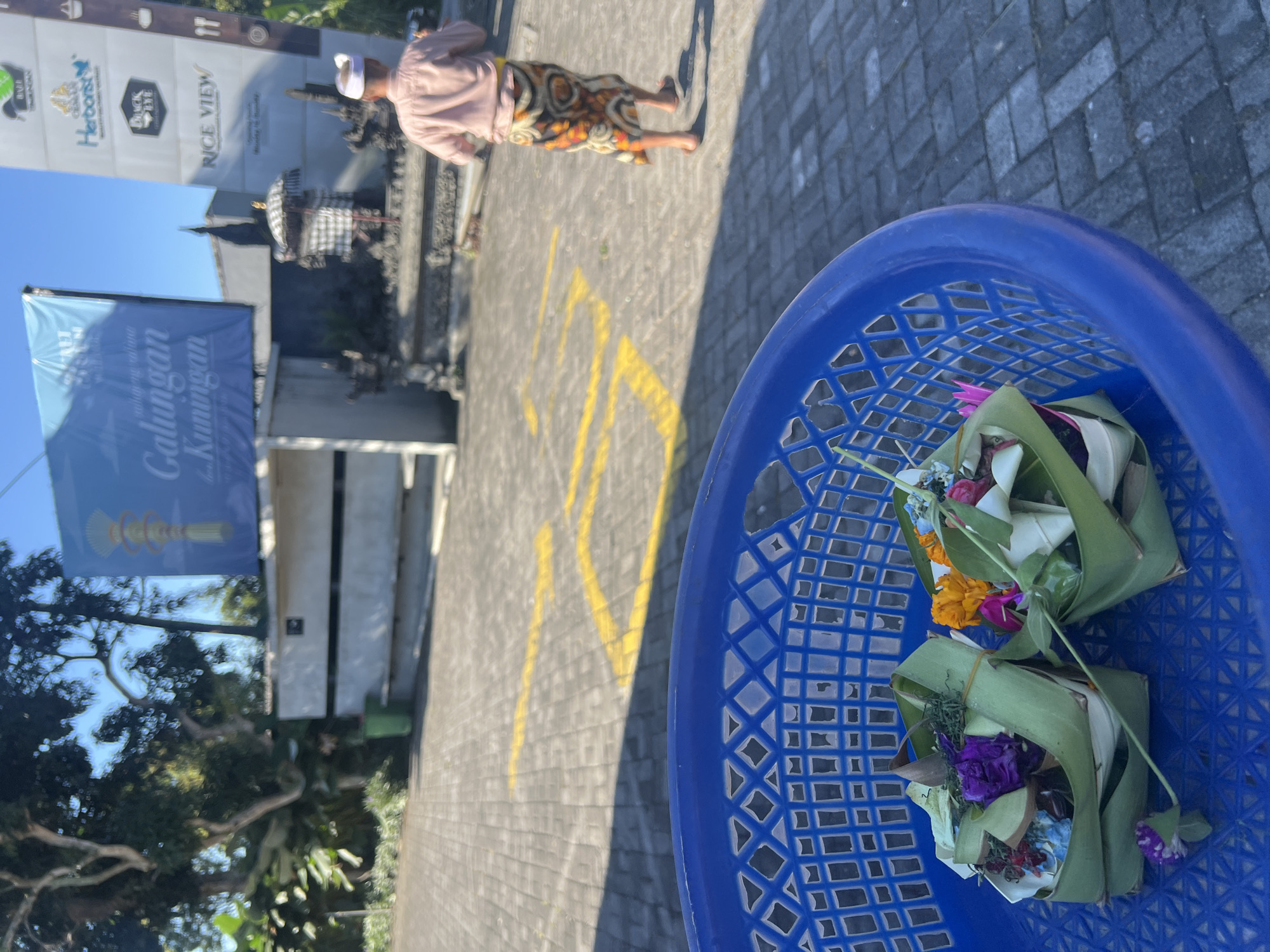 A bright, sunny day in Baturiti, Indonesia is depicted, with a focus on a blue plastic basket in the foreground. The basket contains two small, meticulously crafted offerings made of banana leaves and flowers, indicative of a religious or ceremonial context. In the background, a man dressed in traditional attire, including a sarong and a white cap, walks away from the camera. Behind him, a shrine with an umbrella and intricate stone carvings is visible. A banner with Balinese text and an illustration of a traditional Balinese ornament hangs prominently, suggesting a local festival or event, possibly related to "Galungan dan Kuningan," a significant Balinese Hindu holiday. The ground is paved with interlocking stones, marked with yellow lines, and trees and buildings form the backdrop under a clear blue sky.