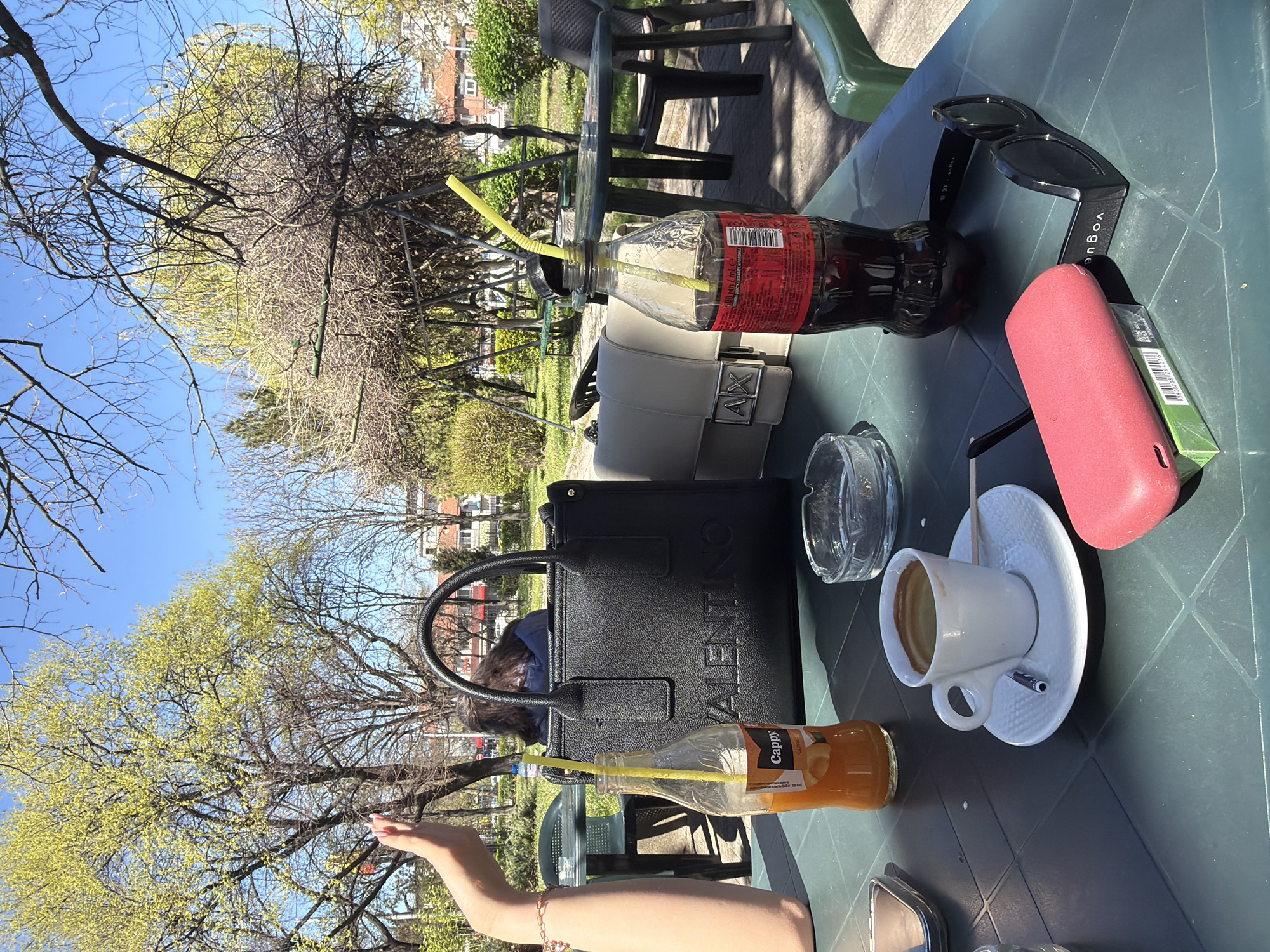 The image shows a table at an outdoor café in Plovdiv, Bulgaria. The table is green and contains a black Valentino bag, a grey Armani Exchange bag, a bottle of cola, a bottle of Cappy orange juice, an ashtray, a cup of coffee on a saucer with a spoon, a red charging case (likely for earbuds), a pack of cigarettes, and a pair of black Vogue sunglasses. A woman is visible in the background. The view behind the table includes trees with budding leaves and buildings, suggesting a park or garden setting within the city. A portion of a woman's arm is also visible, suggesting she is sitting at the table.