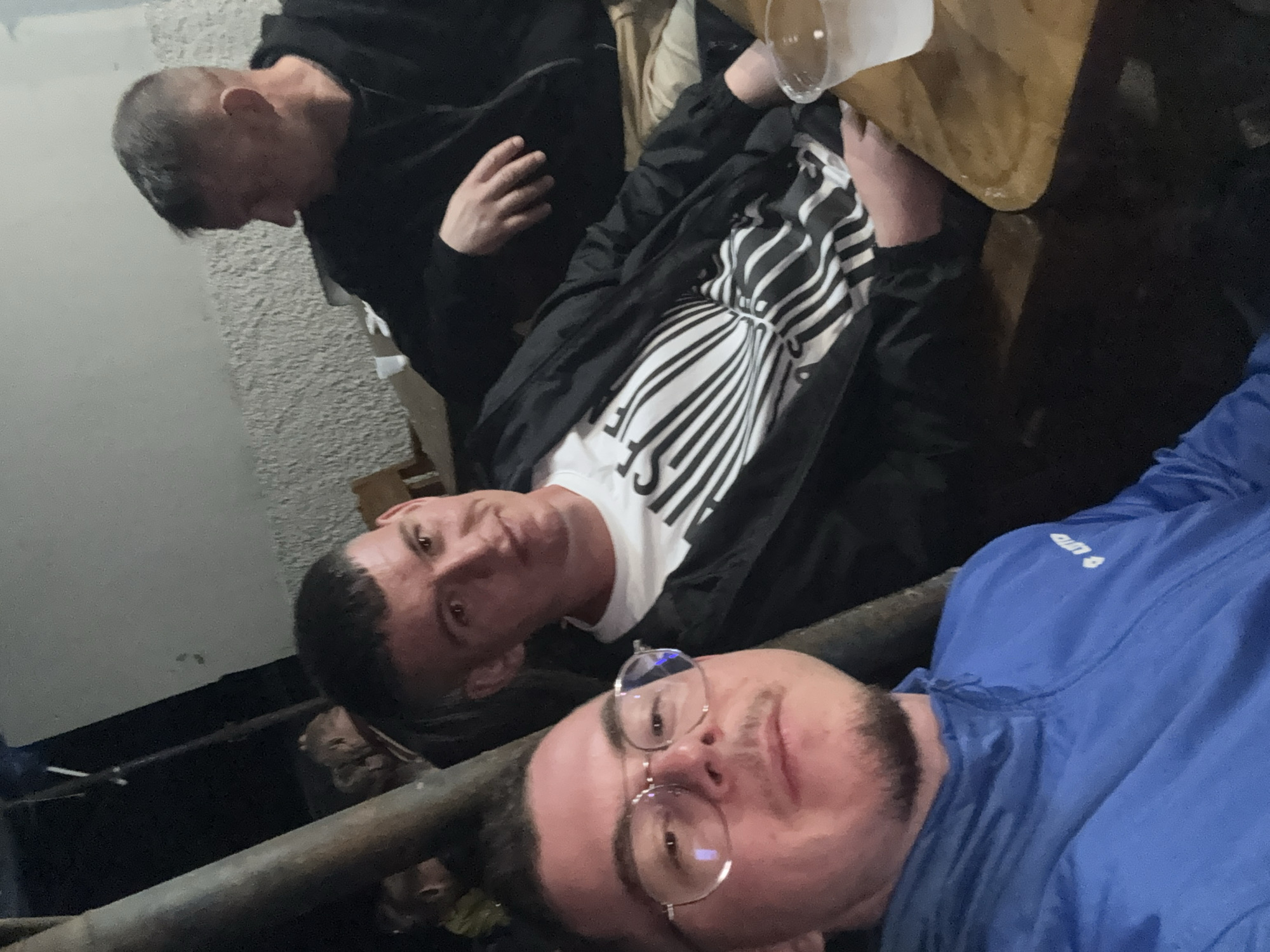 Three men are indoors. One wears a black hoodie, another a black jacket over a white shirt with black lines, and the closest man wears a blue jacket and eyeglasses. The location is potentially Pripek, Bulgaria.