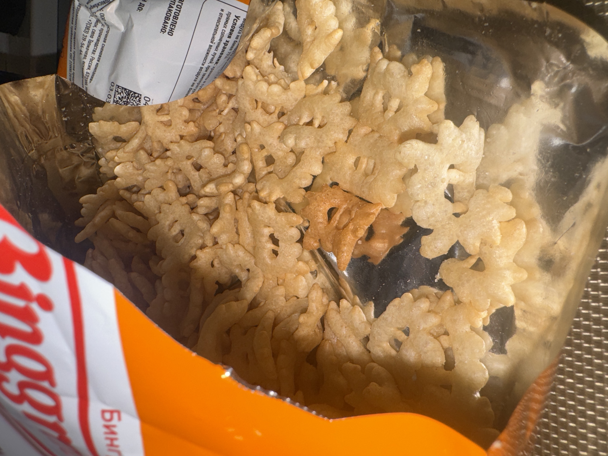 The image shows an open package of Ringg brand snacks, likely potato-based, inside their foil-lined bag. The snacks are light tan, oddly-shaped and partially obscured by the plastic liner. The packaging is predominantly orange, white, and red. Russian text is visible on the packaging and a label attached to the foil liner. Based on the text, the product is manufactured by "Eastern Snack Company" in the village of Tolmachevo, Novosibirsk, Russia. It's unlikely this image shows anything within Krasnoyarsk, as the origin is explicitly stated to be a village in Novosibirsk. The environment includes a patterned surface, which may be a table or countertop.