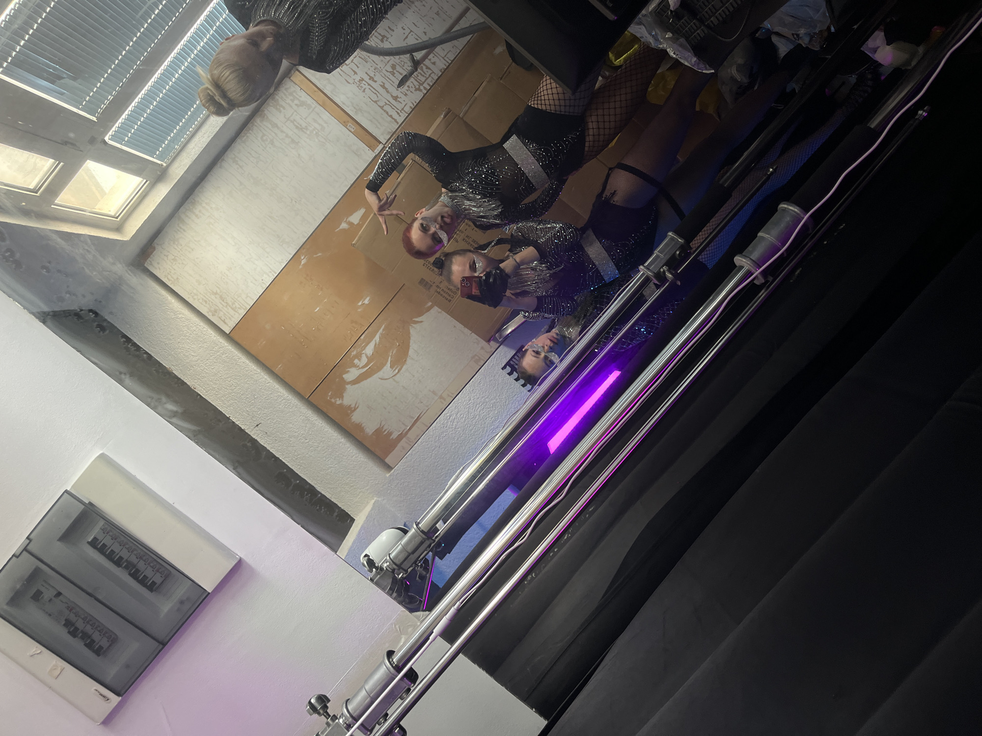 The image depicts a reflective scene, likely within a dressing room or backstage area. Two individuals are reflected in a mirror. The person on the left has red hair and is making a peace sign. Both are wearing similar black outfits with sparkly details. The room itself appears utilitarian, with a large cardboard panel on the wall, a fuse box visible on a white wall to the lower left, and blinds partially covering a window at the top. A lighting rig stands in front of the mirror with a violet tube light illuminated. Black curtains are seen on the right side of the image. The location is reported to be Budva, Montenegro.