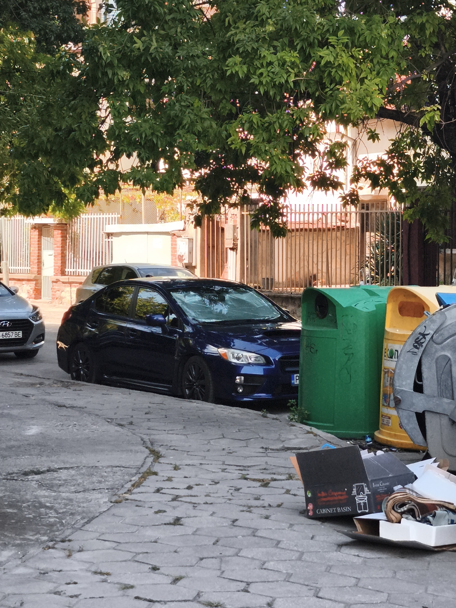 A dark blue Subaru WRX is parked on a cobblestone street in front of a metal fence. The car has black rims and a license plate that reads "PB". To the right of the car are two large plastic trash bins, one green and one yellow. In the foreground, a cardboard box labeled "CABINET BASIN" is on the ground next to some other discarded items. A silver Hyundai Accent is parked to the left of the Subaru. Trees with green leaves are visible in the background, partially obscuring a building. The lighting suggests it is daytime, possibly late afternoon due to the shadows and warm tones.