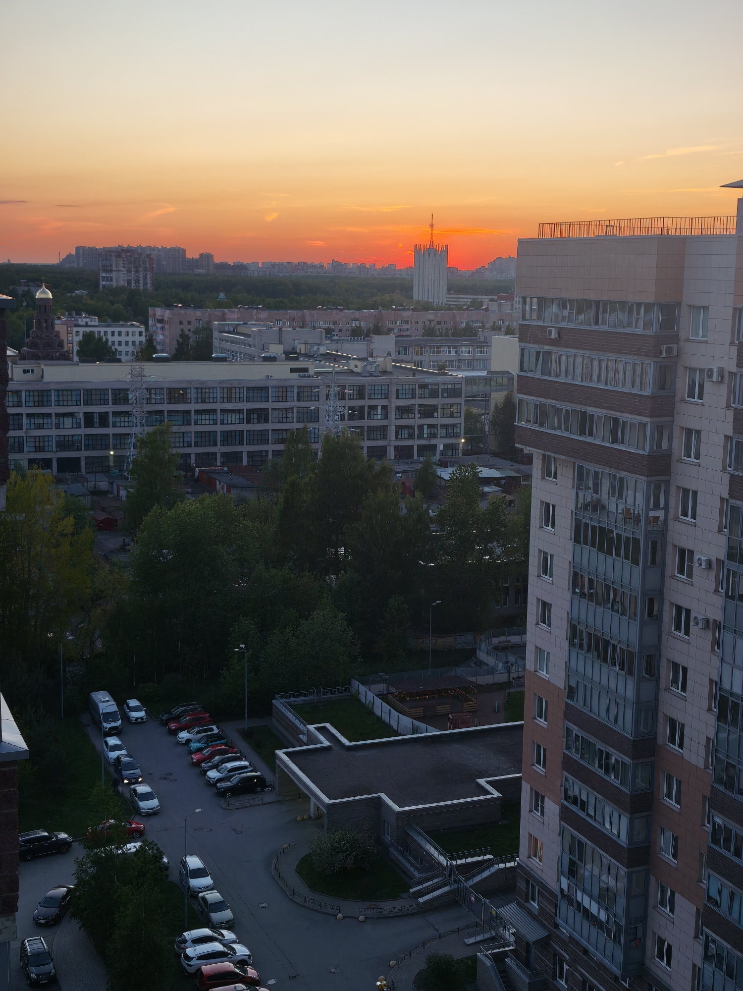 The image depicts a high-angle view of a residential area in Saint Petersburg, Russia, during sunset. In the foreground, there is a tall apartment building with a light brown facade and multiple windows. To the left of the building, there is a parking lot with various cars parked in rows. Beyond the parking lot, there are trees and other buildings, including a large industrial-style building with numerous windows. In the distance, the cityscape is visible, with more buildings and trees. The sky is a vibrant orange and red, indicating sunset. A spire in the distance is the Saint Petersburg TV Tower.
