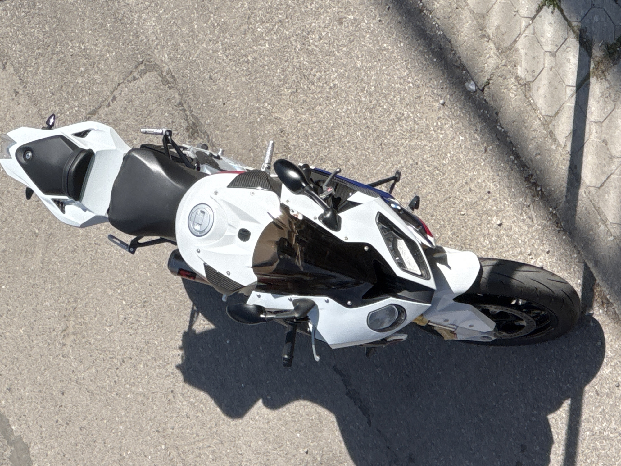 An overhead shot shows a white and black motorcycle parked on asphalt next to a concrete curb. The motorcycle is oriented diagonally across the frame, with its front wheel pointing towards the upper right corner. The setting appears to be an urban environment. There are no people visible in the image. Based on image analysis and context, the location might be in Sofia, Bulgaria.