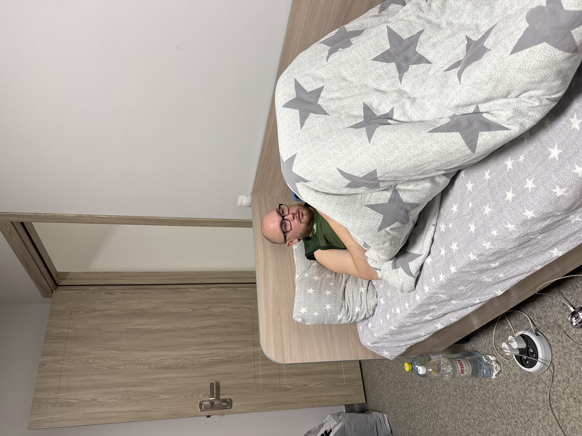 A man with glasses lies in a bed covered with a star-patterned comforter. The bed is located in a room with a wooden cabinet. Next to the bed, on the floor, are bottles of water and a power strip. The bottles are labeled with Cyrillic text, including "ЛИДЖИЙ". Based on the Cyrillic text and the likely name association, the location is probably Kazan, Russia.