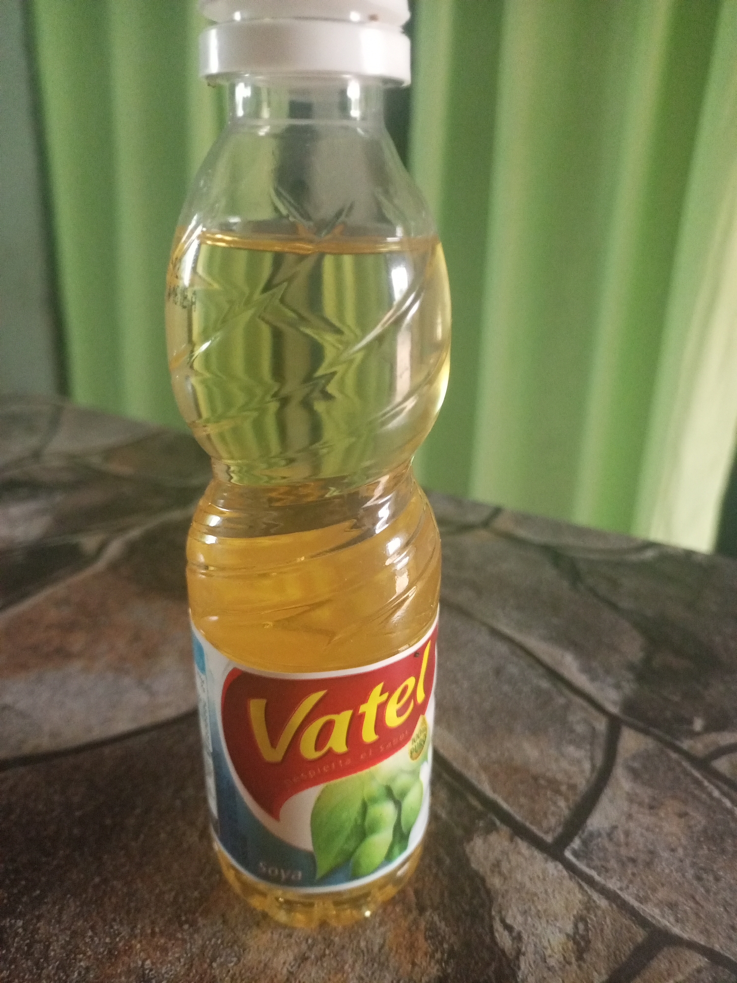 A bottle of Vatel soya oil stands on a brown, faux-stone patterned surface. The bottle is clear plastic with a white cap, and the oil inside is a light yellow color. The label is red, blue, and white, featuring the Vatel logo and an image of soybeans. Behind the bottle is a green, textured curtain. There's no way to verify whether it's in Juan Griego, Venezuela from the image. No people or interactions are visible.