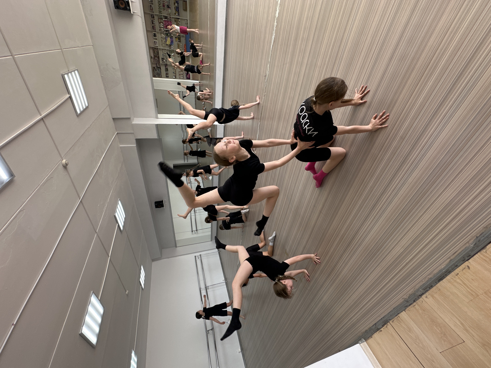 **Scene Description:**
The image captures a dance studio in Votkinsk, Russia, featuring a group of young girls in black leotards practicing ballet. The studio is equipped with a full-length mirror along one wall, reflecting the dancers and the room. The dancers are engaged in various stretching and balance exercises, some using the wall for support. The room is well-lit with rectangular ceiling lights and has a light-toned wooden floor. The studio walls are painted white with paneling details. There is a pin board covered in images and momentos on one side of the mirrored wall.