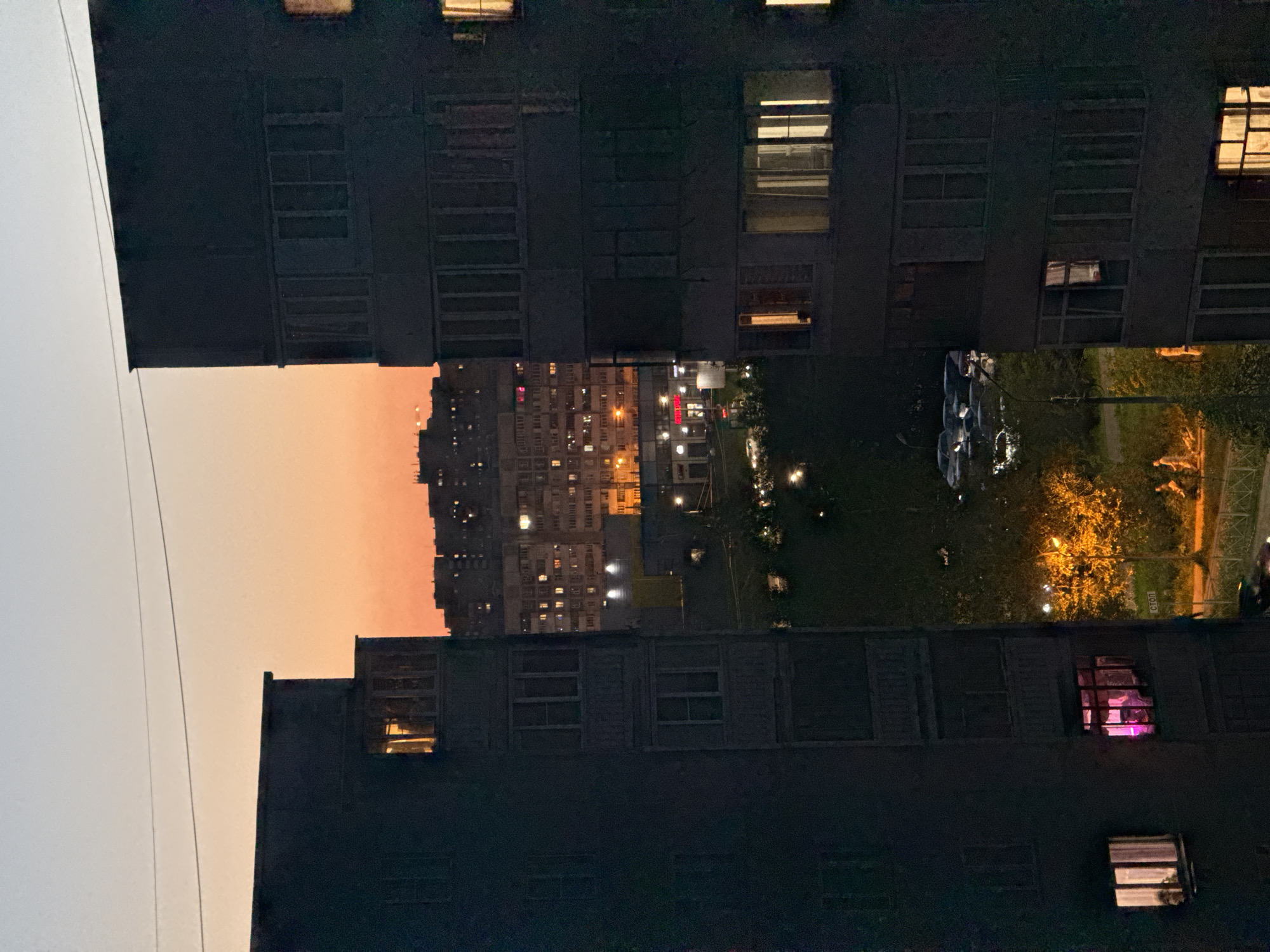 The image shows an urban scene at twilight, taken from a high vantage point. The perspective is slightly tilted.
In the foreground, the dark silhouettes of buildings frame a view into the distance. Windows of these buildings are visible, some illuminated.
In the distance, additional buildings are visible, with numerous lights suggesting they are residential areas. Between the buildings, the sky is visible, displaying shades of orange and pink from the setting sun.
To the right of the distant buildings, a park or green space is illuminated by streetlights. Details such as trees and pathways can be discerned.
Based on the buildings and general environment, the location is possibly Kazan, Russia, but this cannot be determined with absolute certainty.