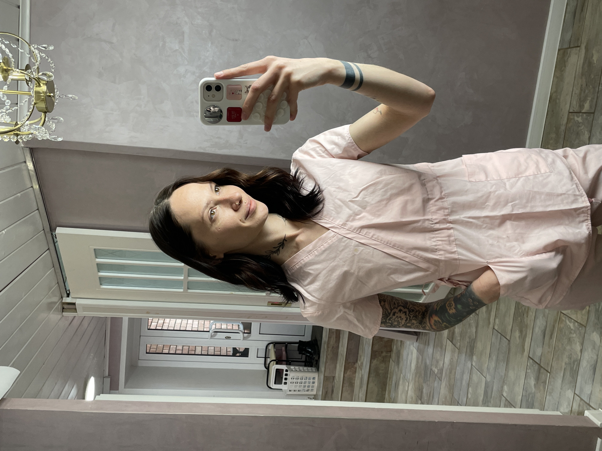 A person takes a selfie in a room. The person is holding a phone up to their face, and wearing a light pink shirt. They have dark hair and tattoos on their arms and neck. The room has light walls and a white door with glass panels. In the background, there is a hallway with a tile floor. A chandelier is partially visible in the upper-left corner. It is not possible to confirm the location in the city of Izhevsk, Russia from the image.