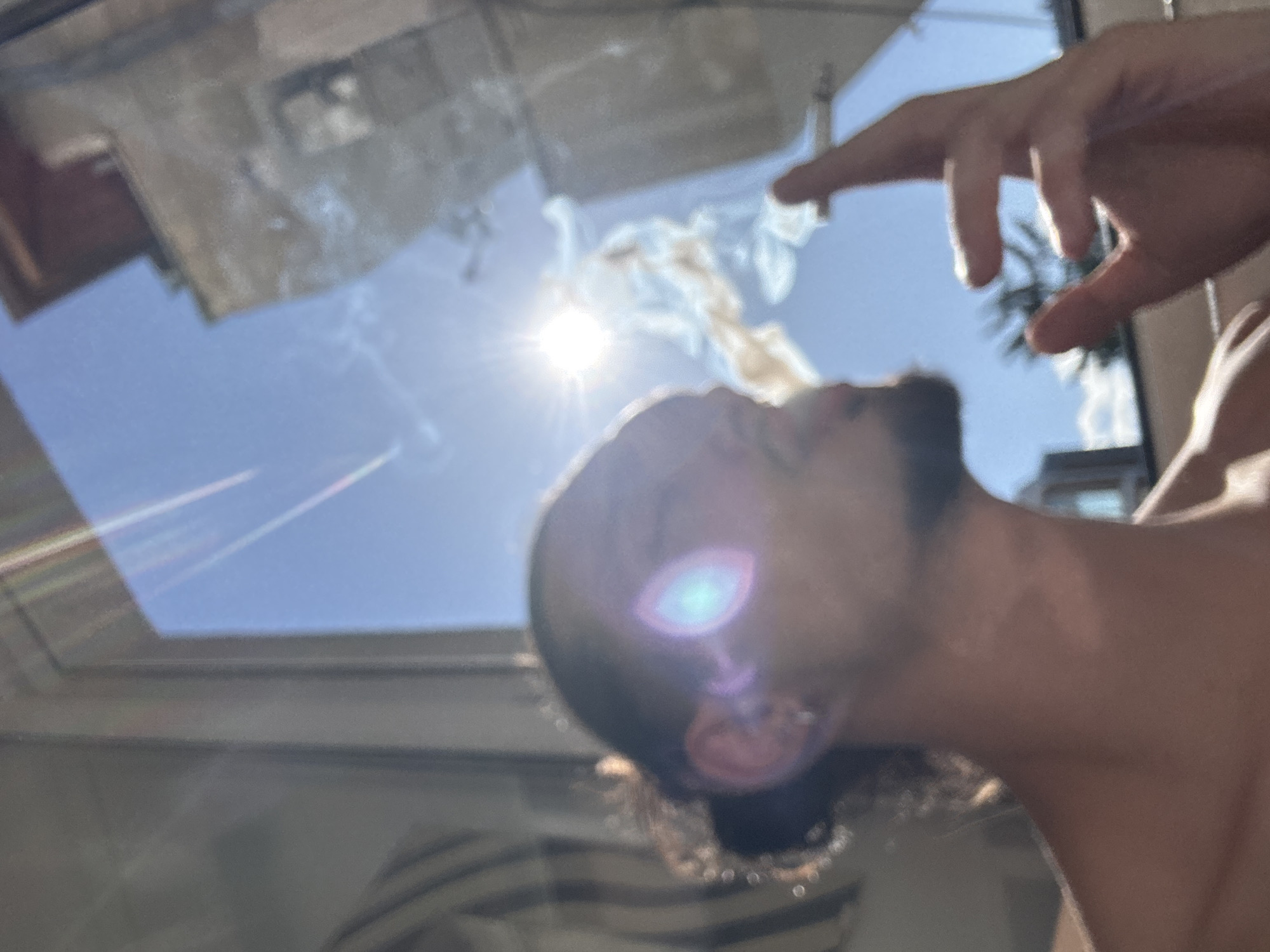 The image shows a person smoking outdoors, with a bright sun in the background. The person is seen in profile, with their head turned to the right. They have dark hair tied back in a bun and are wearing an earring. Smoke is emanating from the cigarette held in their right hand. The background consists of a clear blue sky, a building, and what appears to be a window frame. The overall lighting suggests it is daytime, with the sun being a prominent element. The scene is likely set in an urban environment, possibly a balcony or window overlooking a city, but no specific location in Sofia, Bulgaria can be identified from the visual information provided.