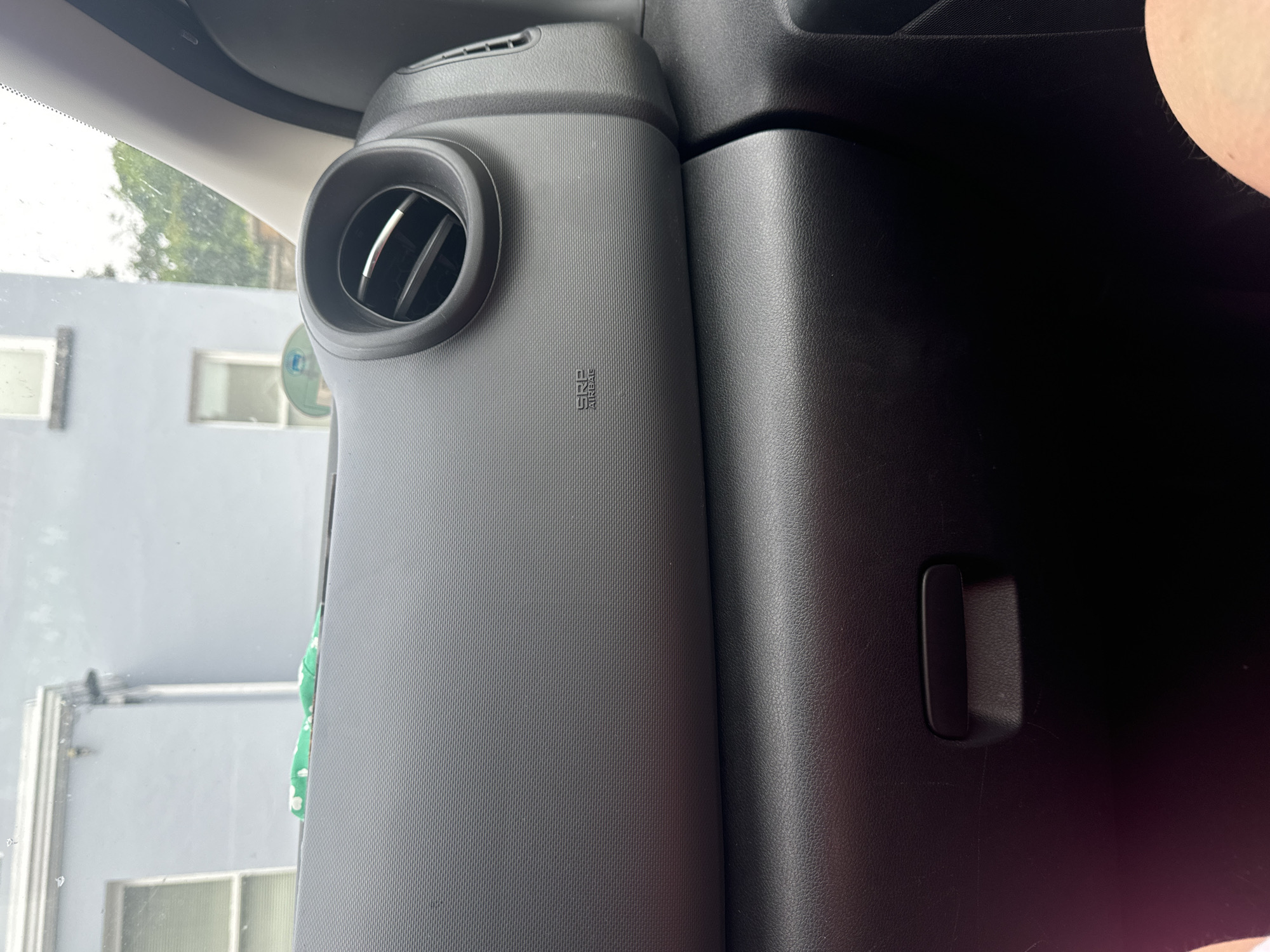 The image shows the interior of a car. In the foreground, the dashboard is visible, with a textured black surface. A circular air vent is located on the right side of the dashboard. Below the vent, a small black emblem with the words "SRS AIRBAG" is imprinted on the dashboard. A black glove compartment is visible below the dashboard. In the background, through the car's windshield, a light blue building with white-framed windows can be seen, along with some green trees and foliage. The scene appears to be in an urban or suburban setting. The image was taken on a cloudy day. The image is a close-up shot of the car's interior and the exterior view is partially obscured. There are no people clearly visible in the image.